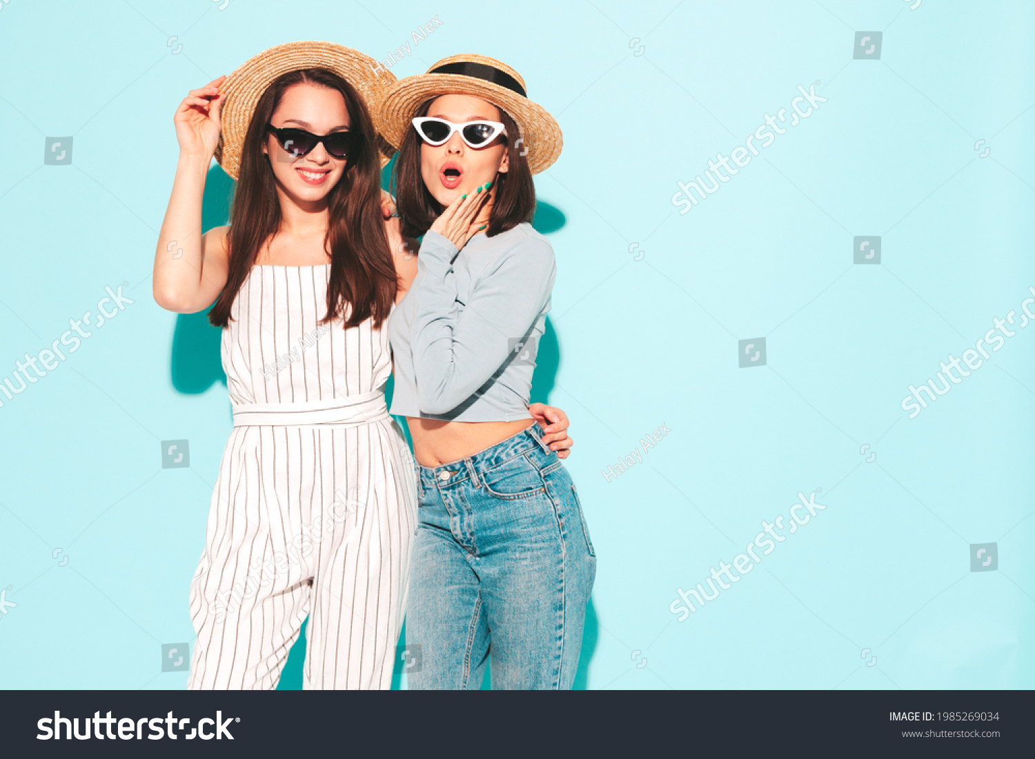 Two young beautiful smiling hipster female in trendy summer  clothes.Sexy carefree women posing near light blue wall in studio.Positive and cheerful models in hats #1985269034