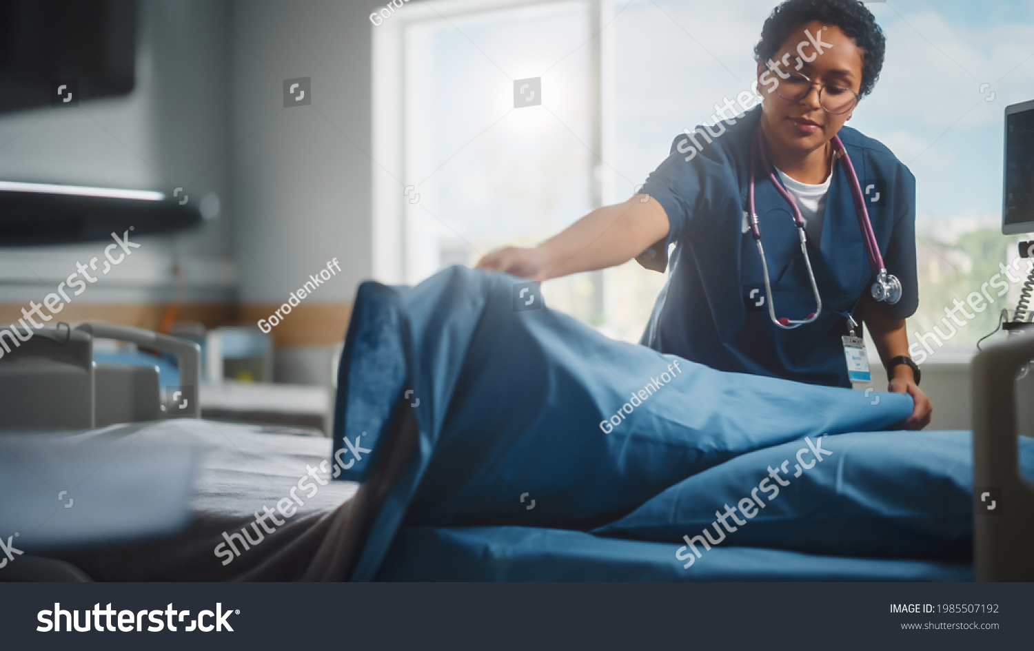 Hospital Ward: Beautiful, Professional Latin Black Nurse Working, Doing Bed, Cleaning Room After Happy Healthy Patients Recover and are Discharged Under Treatment of the Best Doctors. Sunny Room #1985507192