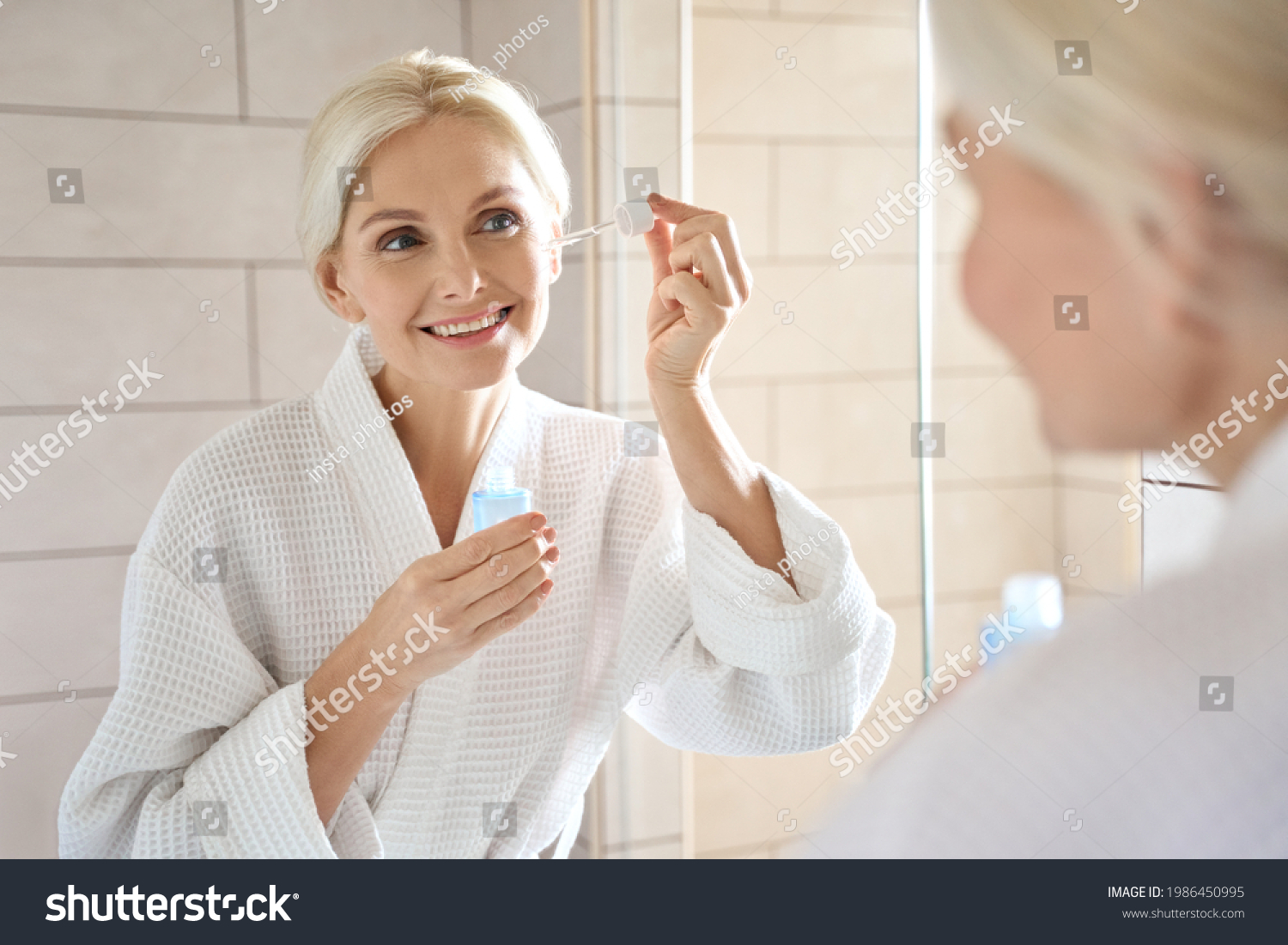 Senior mature older caucasian woman touching clean face eye contour with antiaging pipette serum essence oil looking at mirror wearing bathrobe. Anti wrinkle prevention skin care products concept. #1986450995