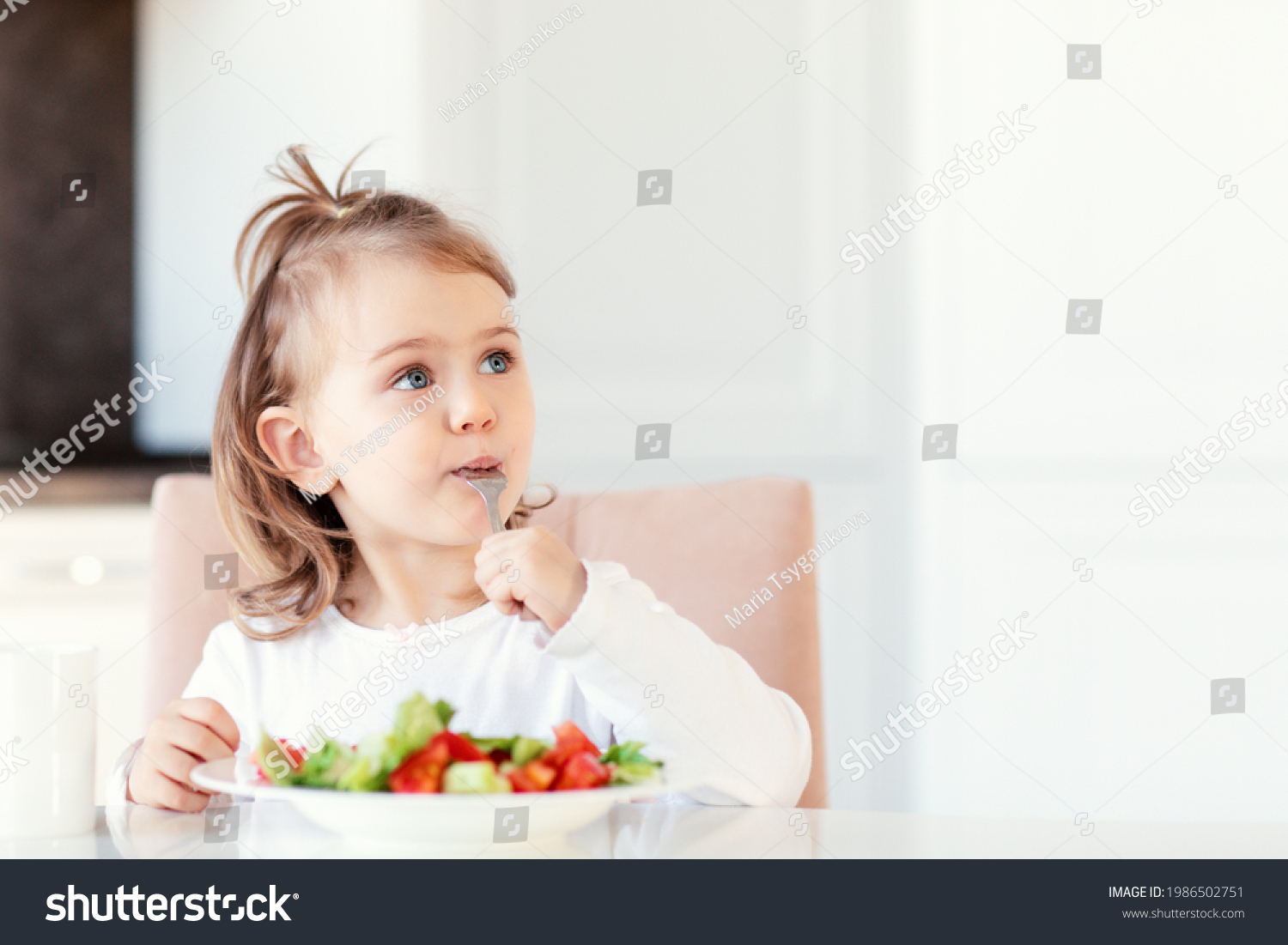 Child girl eating fresh raw vegetables vitamin salad in white kitchen background and thoughtfully looks at the empty copy space.Healthy nutrition food for children. Place for text for advertising. #1986502751