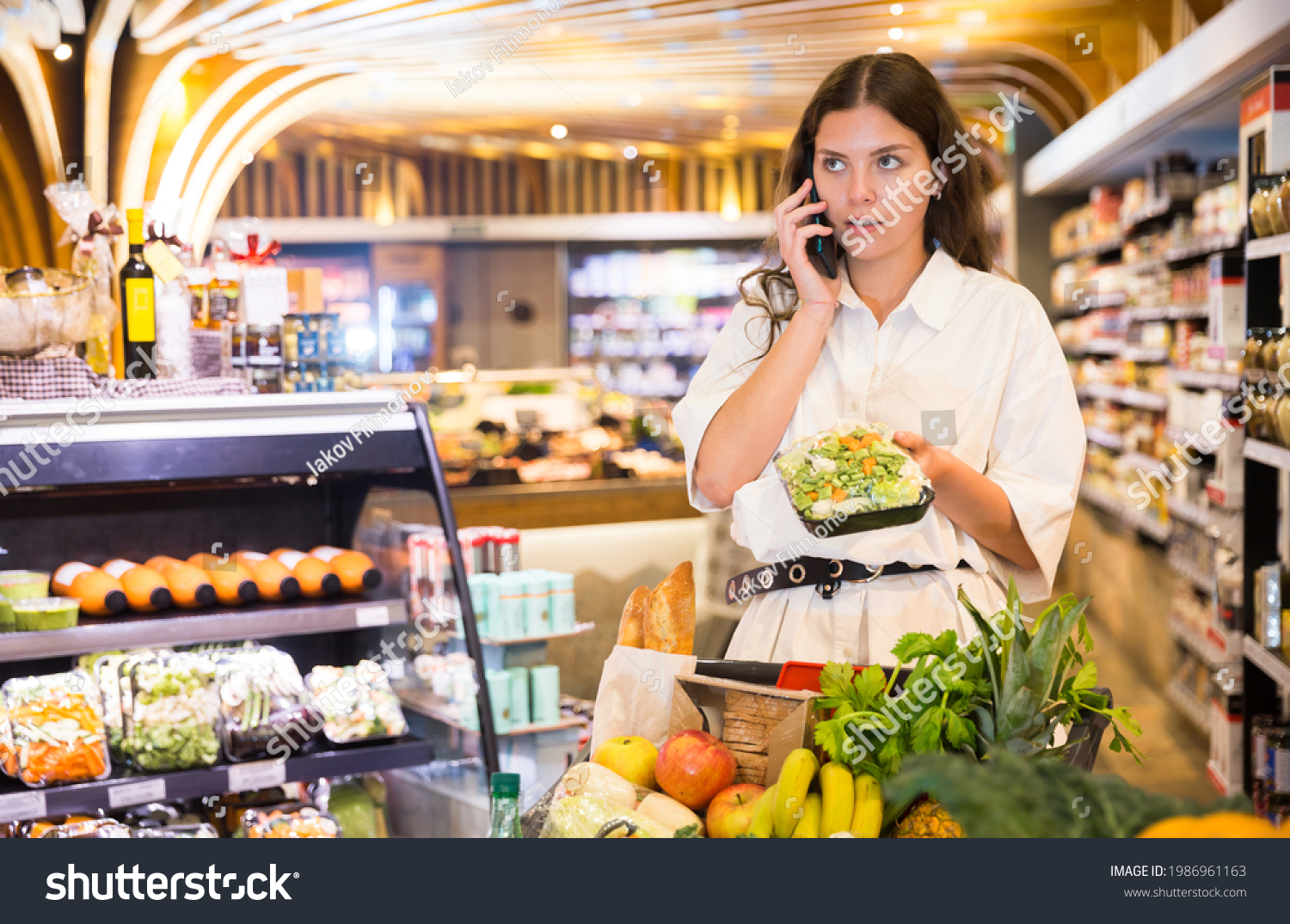 Confident girl in a supermarket is talking on a mobile phone, choosing frozen vegetables on a in the healthy food..department #1986961163