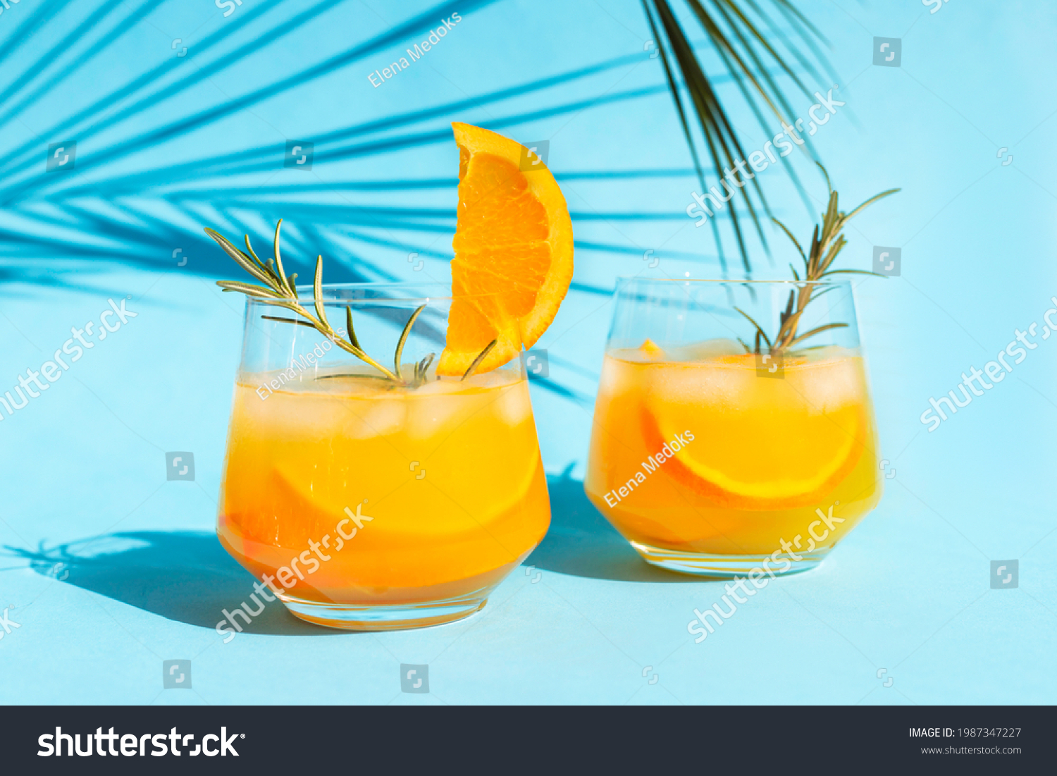 Close-up lemonade, orange cocktail, refreshing summer drink with ice in a glass with a sprig of rosemary on a blue background under a palm leaf on a bright sunny day. #1987347227