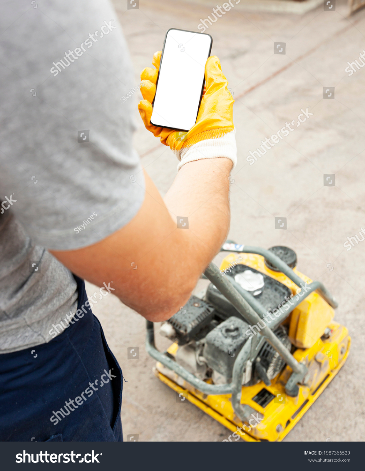 The worker holds the phone while tamping a gravel by the vibration plate. Mockup for house repair or building #1987366529