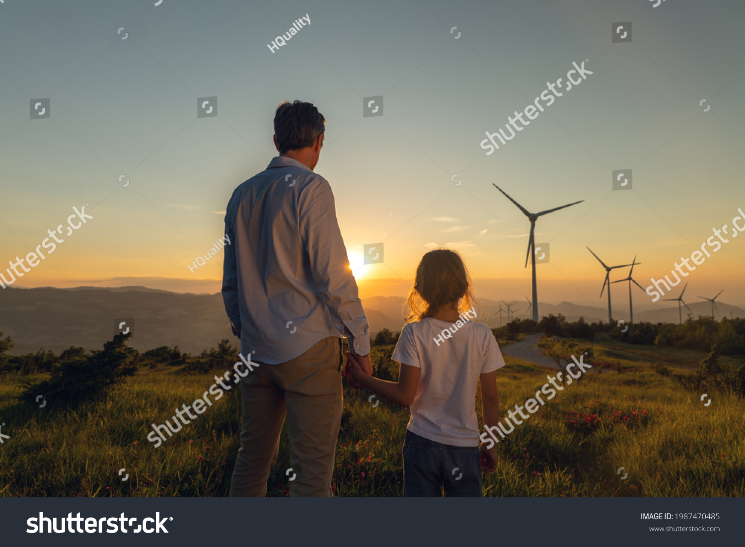 Cinematic shot of carefree young father engineer keeping his daughter for hand and looking on windmill field at sunset. Concept of renewable energy, love for nature, family, electricity, green, future #1987470485