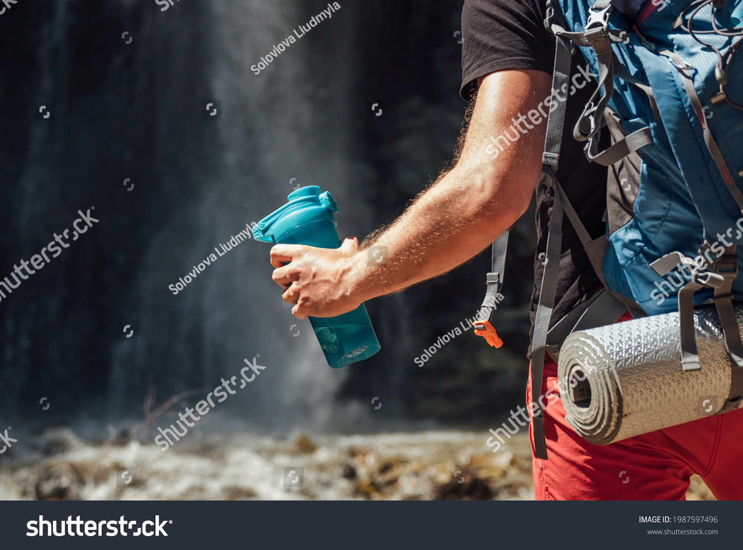 Hand close up with drinking water bottle. Man with backpack dressed in active trekking clothes touristic staying near mountain river waterfall and enjoying Nature. Traveling, trekking concept image #1987597496