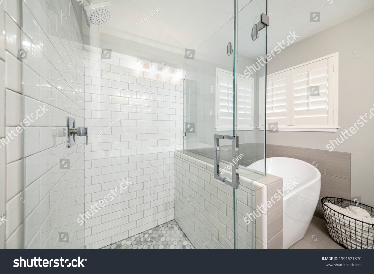 Bathroom interior with frameless shower stall and freestanding bathtub. Stall with subway tile walls and wall mounted shower head beside the tub with towel storage basket against the jalousie windows. #1991621870