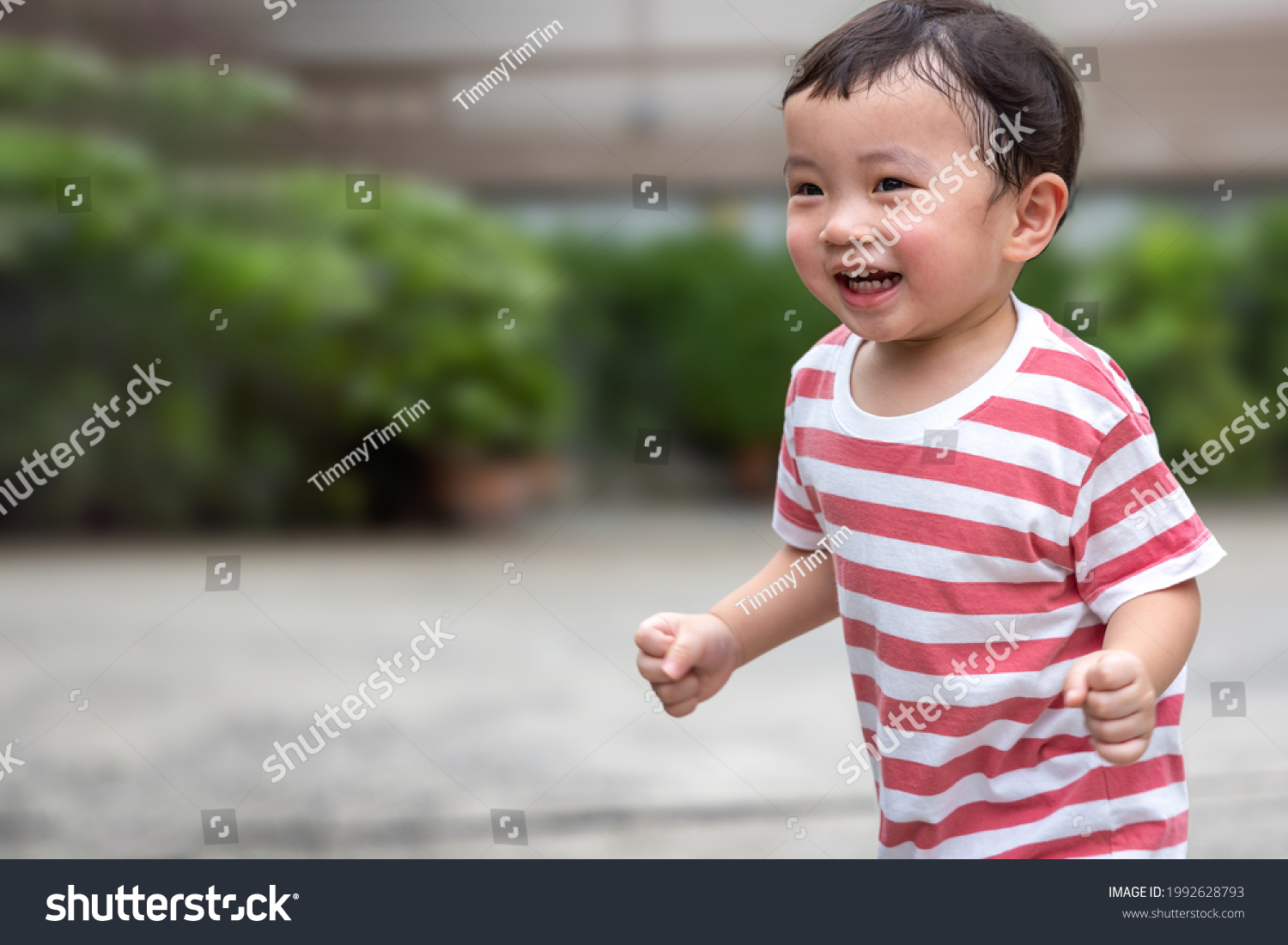 Portrait of an asian boy (toddler) running toward and smiling with happy and fun face while playing outdoor. A Child wear striped shirt in red and white color. Head and hair is wet by sweat. #1992628793