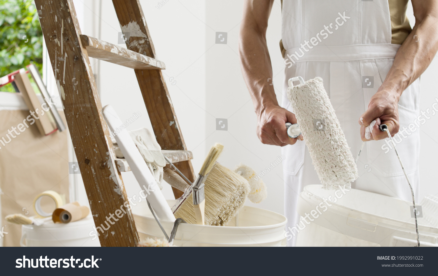 hands of house painter man decorator work of home to renovate, using roller paint and holding white bucket, a wooden ladder with paint brushes as background, close-up #1992991022