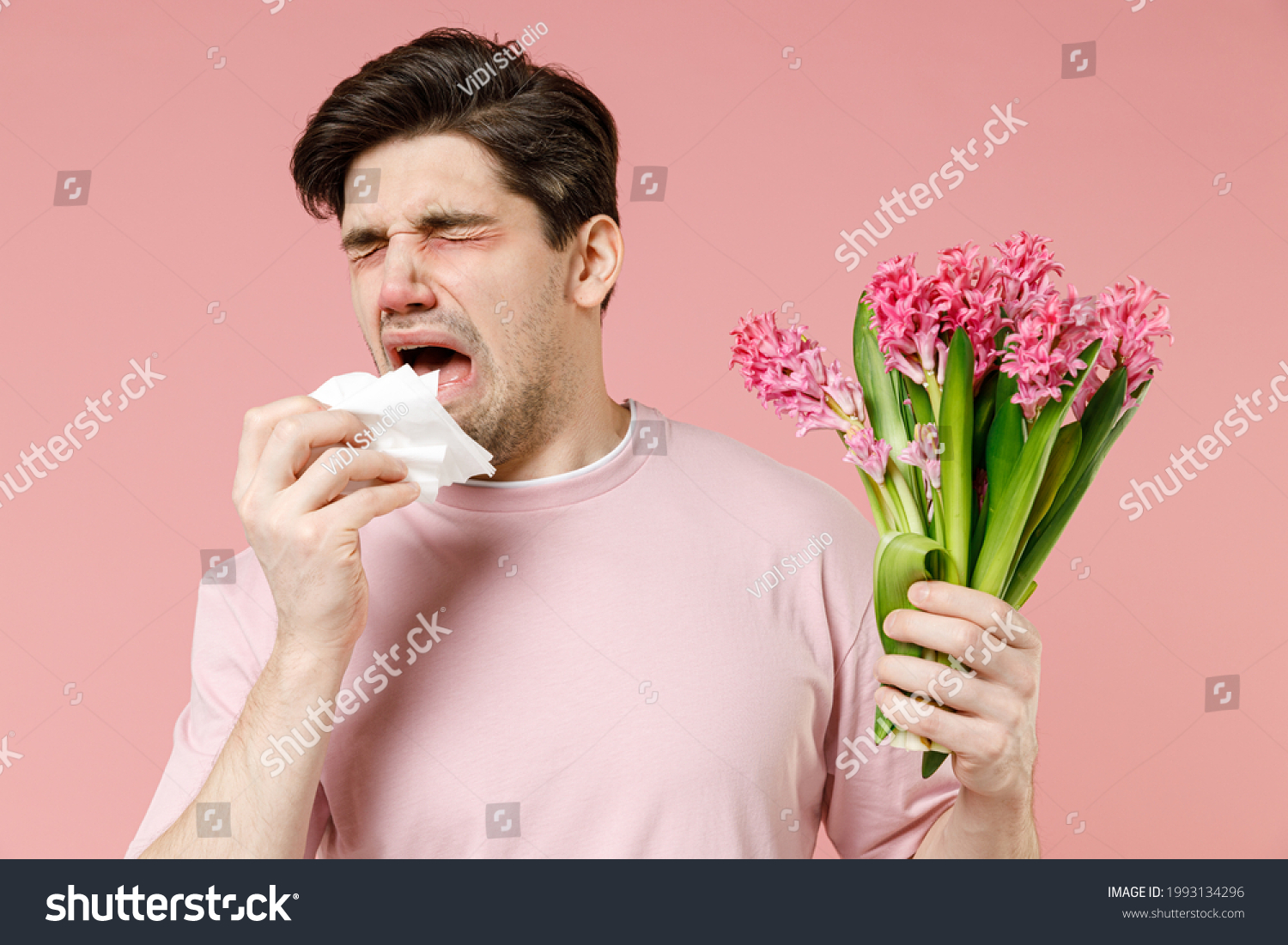 Sick allergic man has red eyes runny stuffy sore nose suffer from pollen allergy symptoms hay fever hold bloom flower plant napkin reaction on trigger isolated on pastel pink color background studio #1993134296