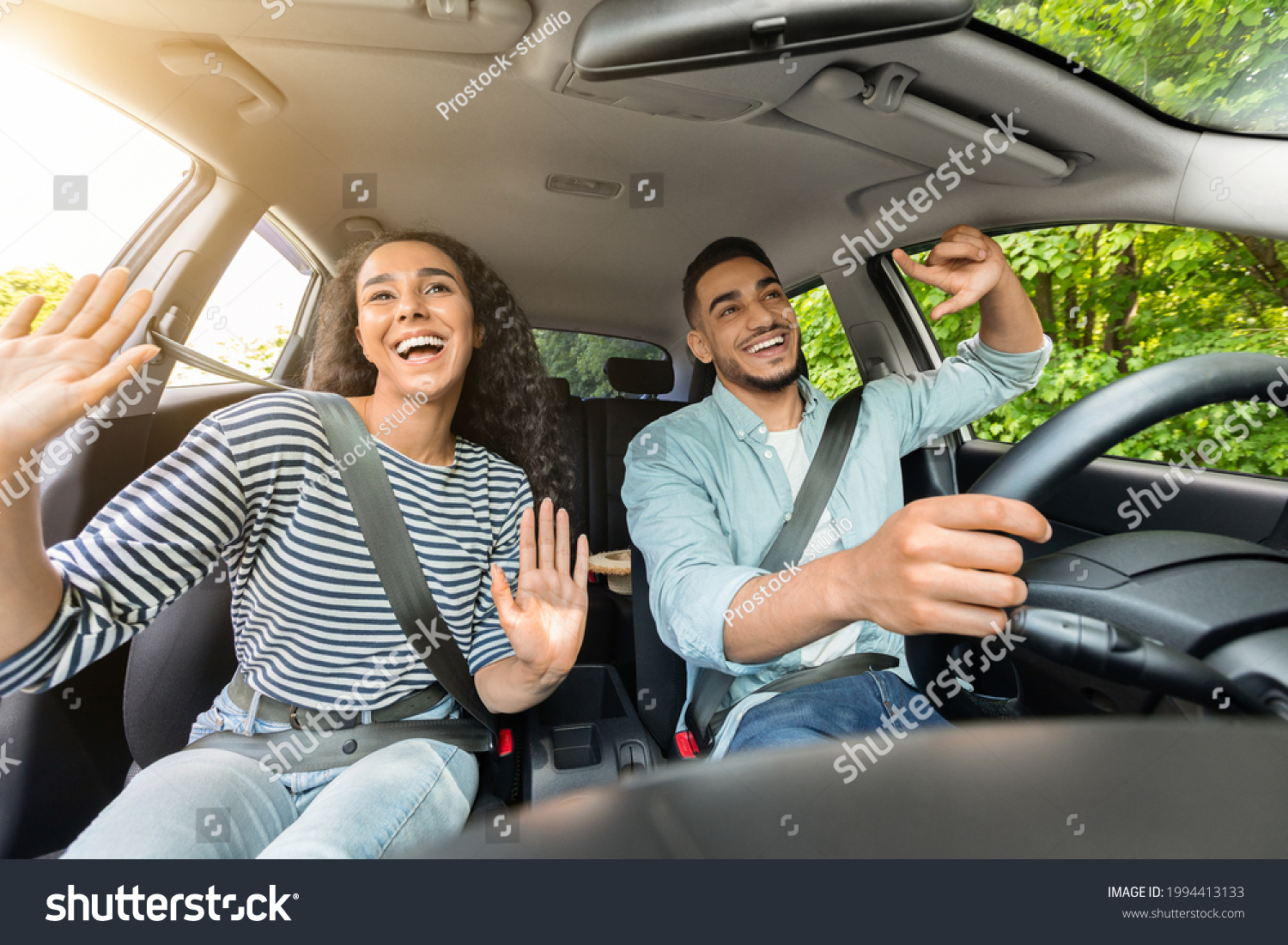 Emotional millennial mixed race couple handsome arab guy and pretty brunette lady having fun while car trip, listening to music and cherfully singing, shot from inside the auto, sun flare #1994413133