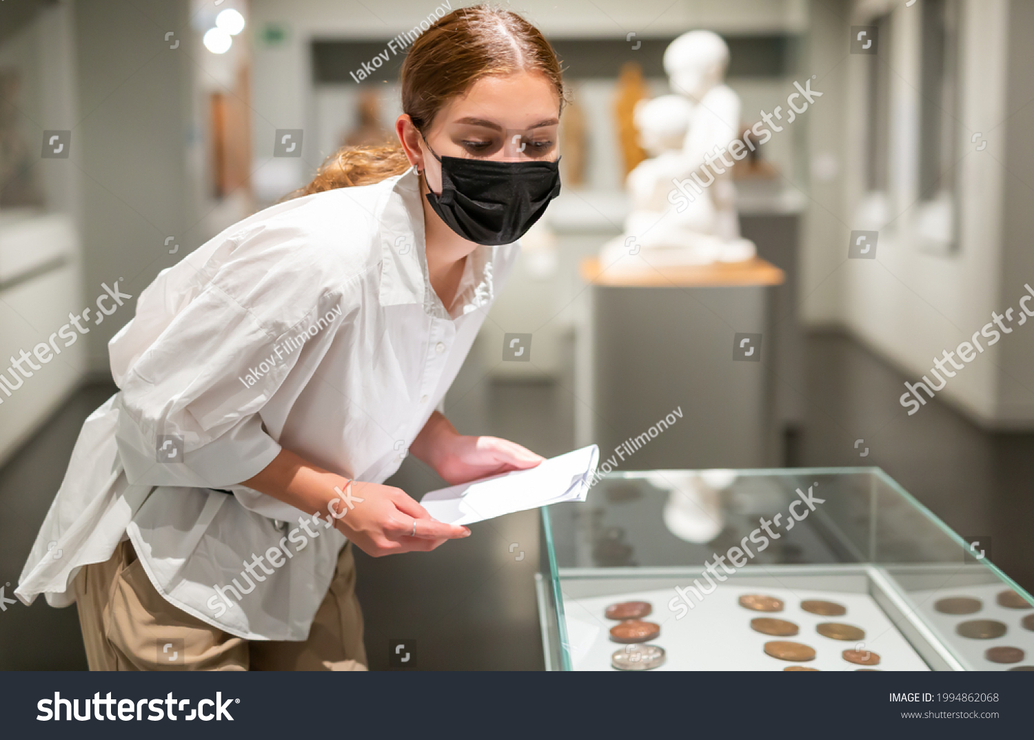 Young positive female in protective face mask with information brochure visiting museum and looking at the exposition of ancient coins #1994862068
