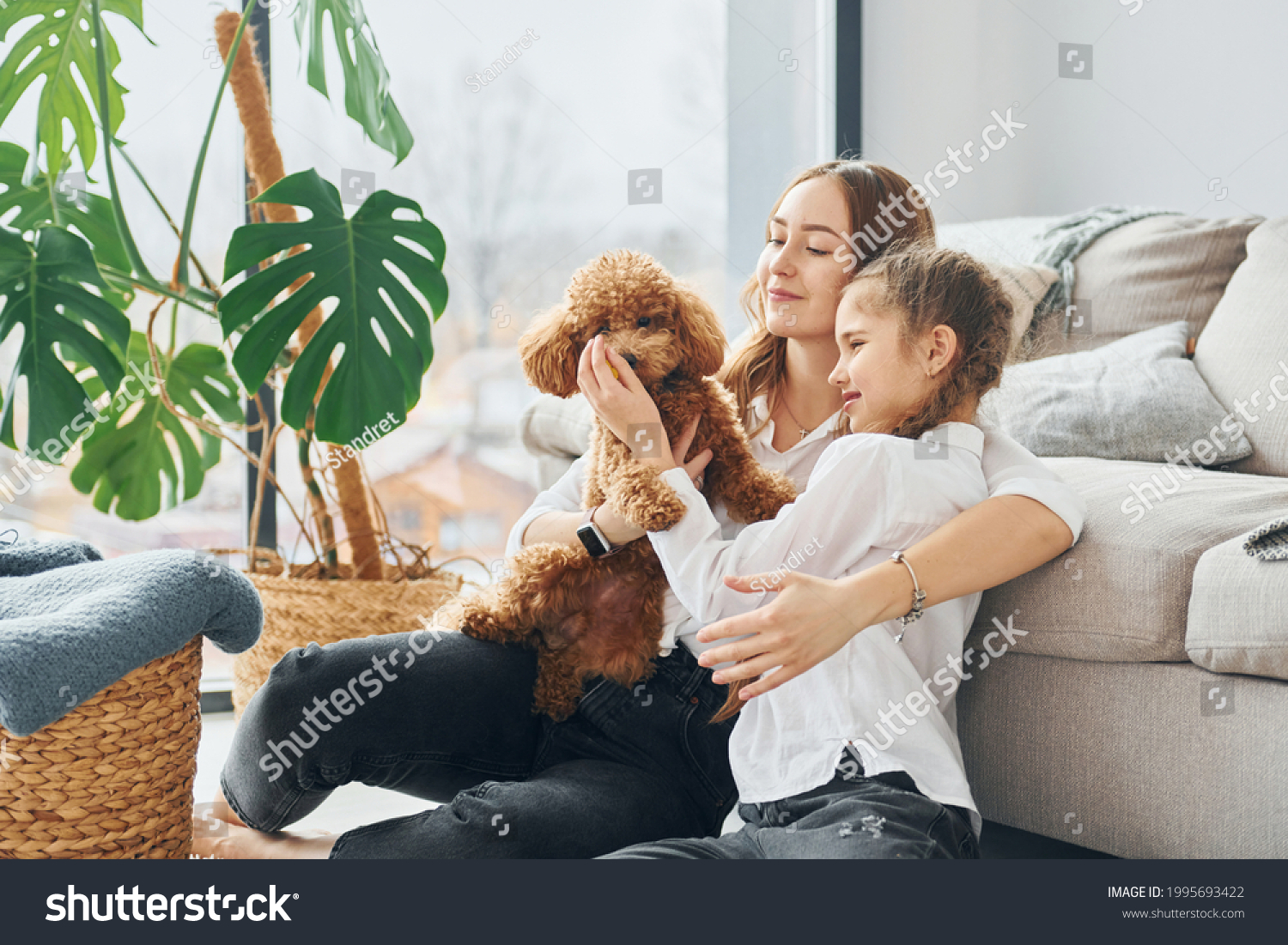 Mother with her daughter playing with dog. Cute little poodle puppy is indoors in the modern domestic room. #1995693422