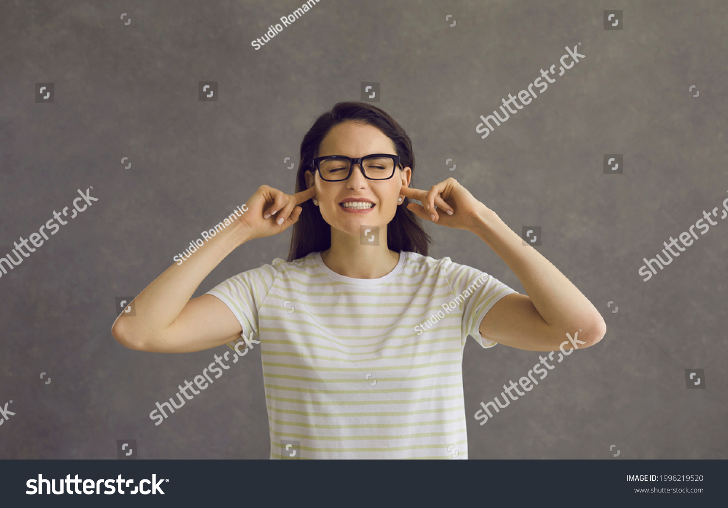I can't hear you. Head shot of young woman in glasses closing eyes and plugging ears with fingers to ignore loud noise, unwanted opinion, dumb stupid comment or useless unsolicited uninvited criticism #1996219520