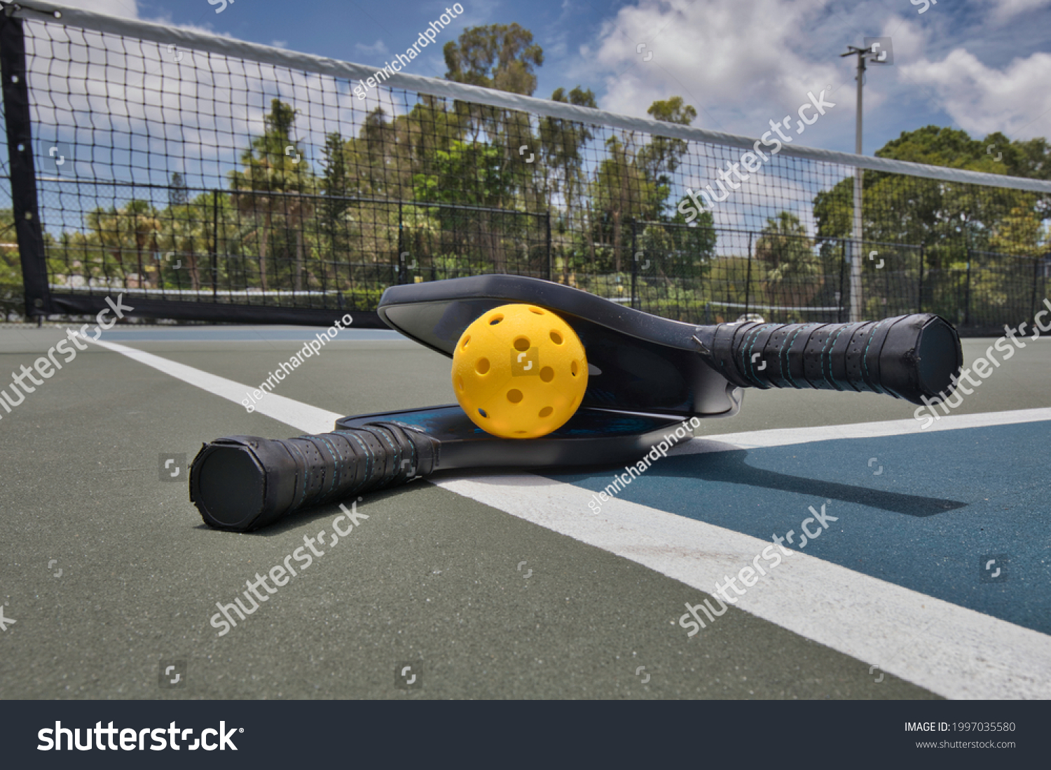 Closeup of a pickleball and paddles on an empty court under sunny skies. #1997035580
