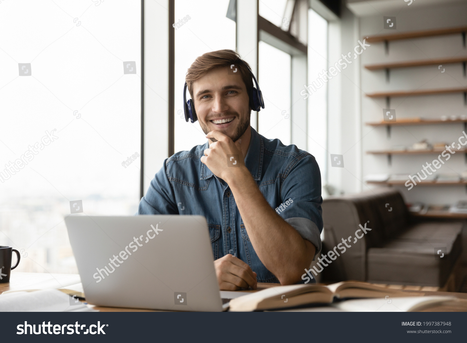 Portrait of happy millennial generation man in headphones sitting at table with books and computer. Smiling young male student posing in modern home office, e-learning distantly on online courses. #1997387948