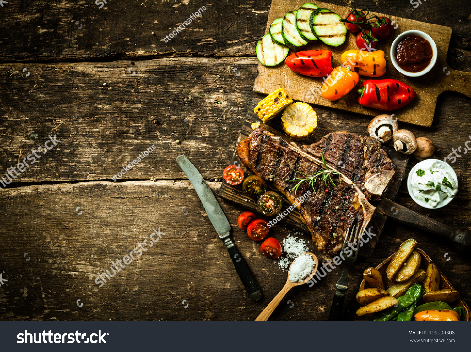 Overhead view of colorful roast vegetables, savory sauces and salt served with grilled t-bone steak on a rustic wooden counter in a country steakhouse #199904306