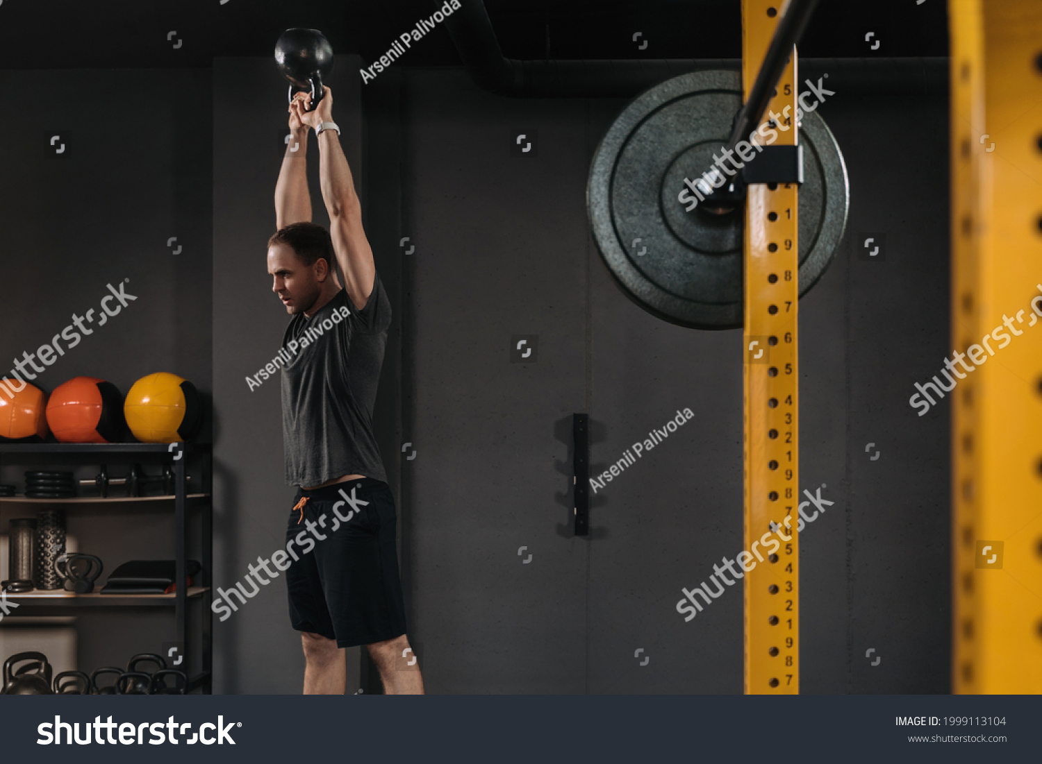 Muscular weightlifter doing an exercise with kettlebell at dark crossfit gym with professional sport equipment. Barbell on front ground. Crossfit athlete having workout, functional training indoors #1999113104