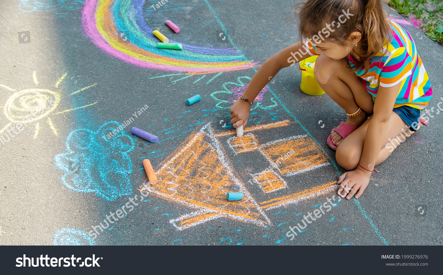 The child draws a house and a rainbow on the asphalt with chalk. Selective focus. Kids. #1999276976
