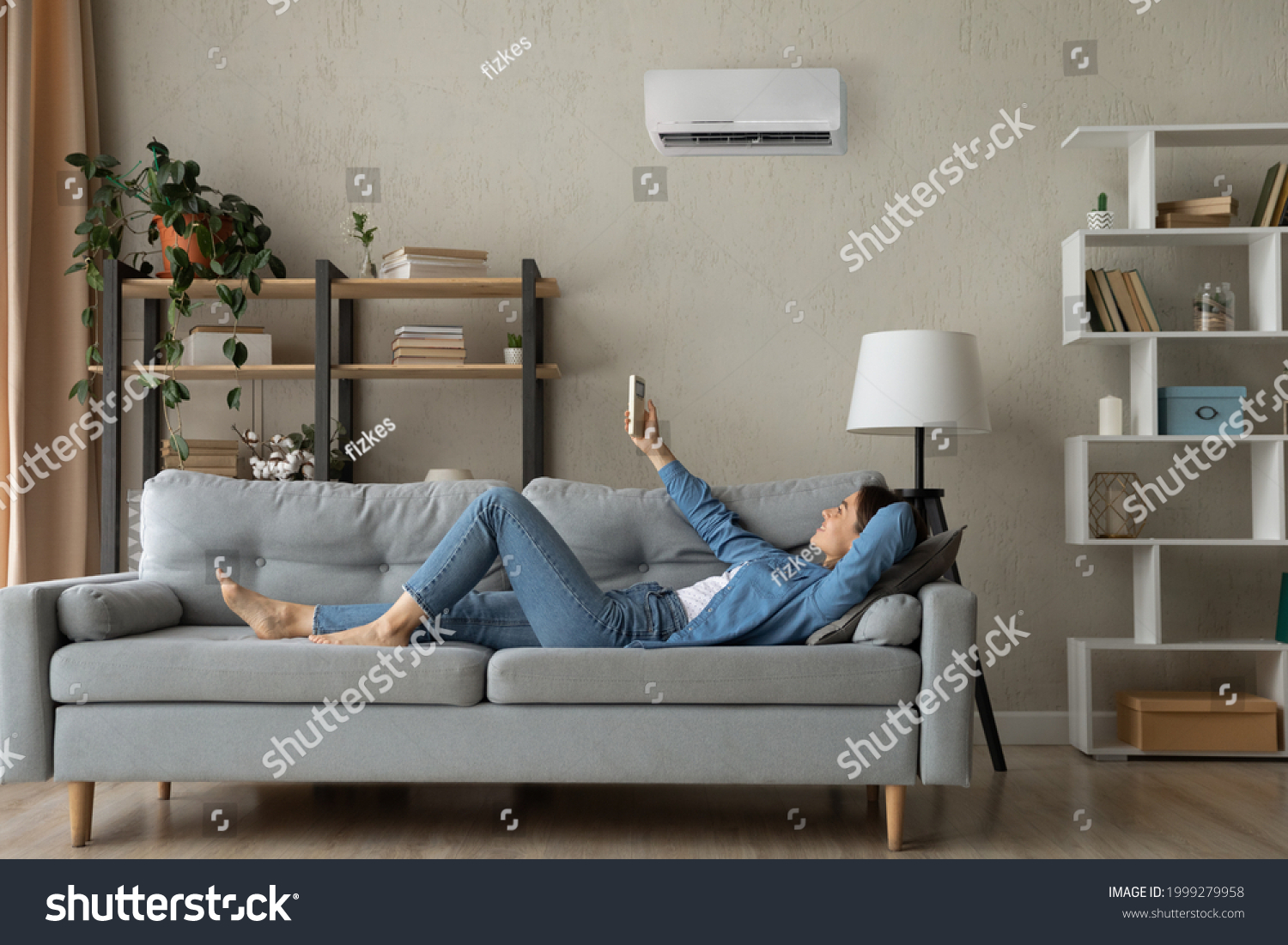 Full length relaxed young happy woman homeowner lying on comfortable sofa turning on air conditioner with remote controller, breathing fresh air, enjoying lazy weekend time, meditating in living room. #1999279958