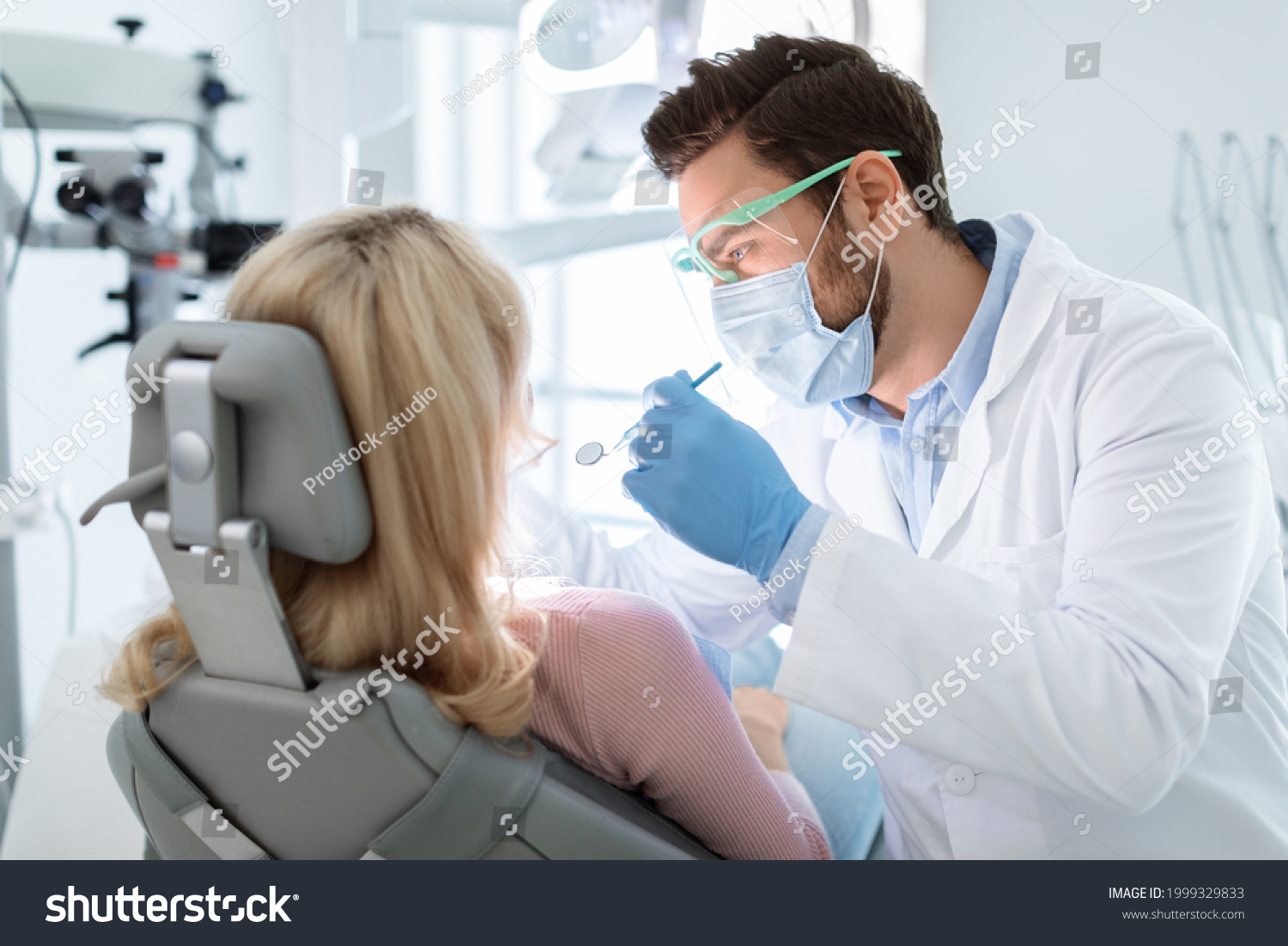 Man dentist in face mask and glasses doing treatment for patient blonde lady, holding dental tools, wearing rubber gloves. Stomatology, dentistry, modern dental clinic concept #1999329833