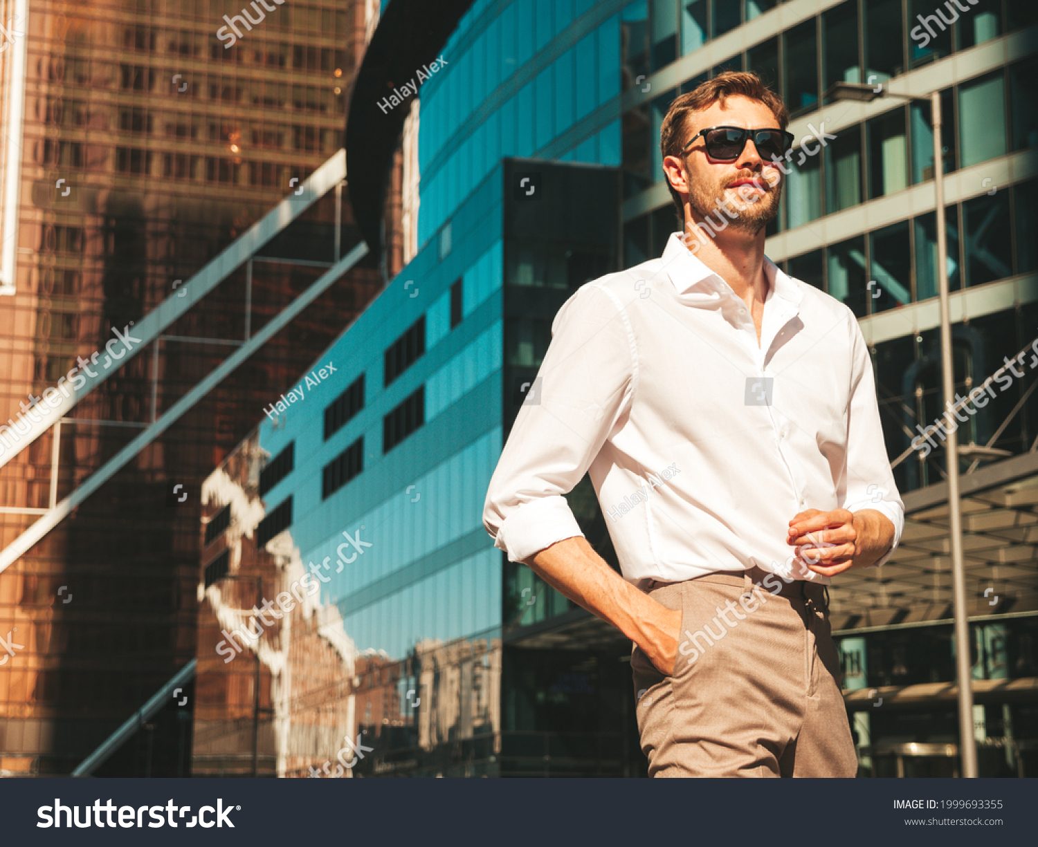 Portrait of handsome confident stylish hipster lambersexual model.Modern man dressed in white shirt. Fashion male posing in the street background near skyscrapers in sunglasses. Outdoors at sunset  #1999693355