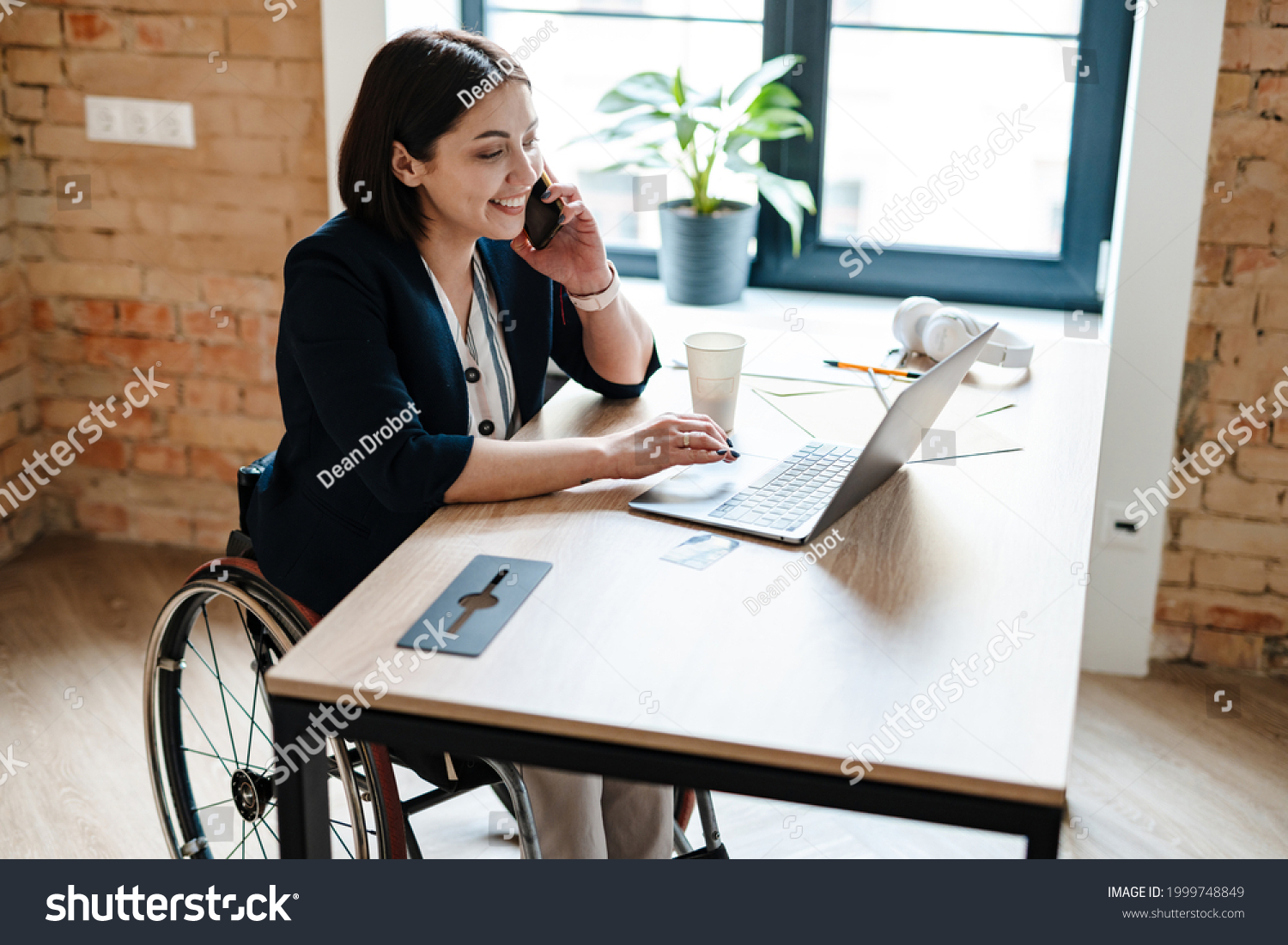Young disabled business woman in wheelchair working at office desk and with laptop, talking on mobile phone accessibility and independence concept #1999748849