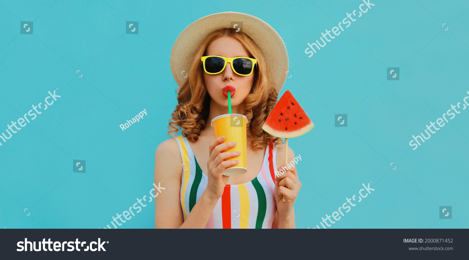 Summer fashionable colorful portrait of stylish young woman drinking juice with lollipop or ice cream shaped slice of watermelon wearing a straw hat on blue background #2000871452