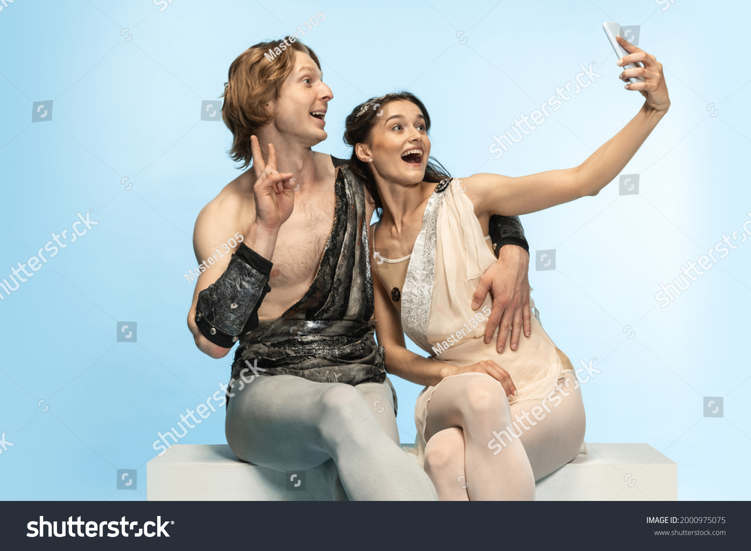 Selfie on mobile phone. Young couple of ballet dancers in ancient Rome costums at blue studio as modern man, woman. Historical character, creative, classical art, humor and comparison of eras concept. #2000975075