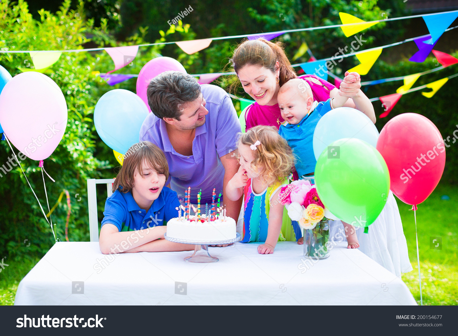 Happy big family with three kids - school age boy, toddler girl and a little baby enjoying birthday party with a cake blowing candles in a summer garden decorated with balloons and banners #200154677