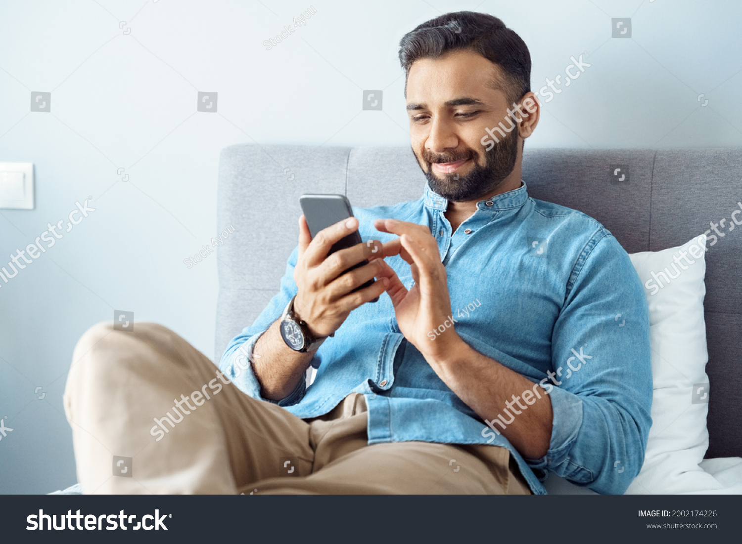 Smiling young adult indian freelance business man using mobile phone checking social media network feed or message chat sitting on bed at home. Online digital communication, rest after hard work day #2002174226