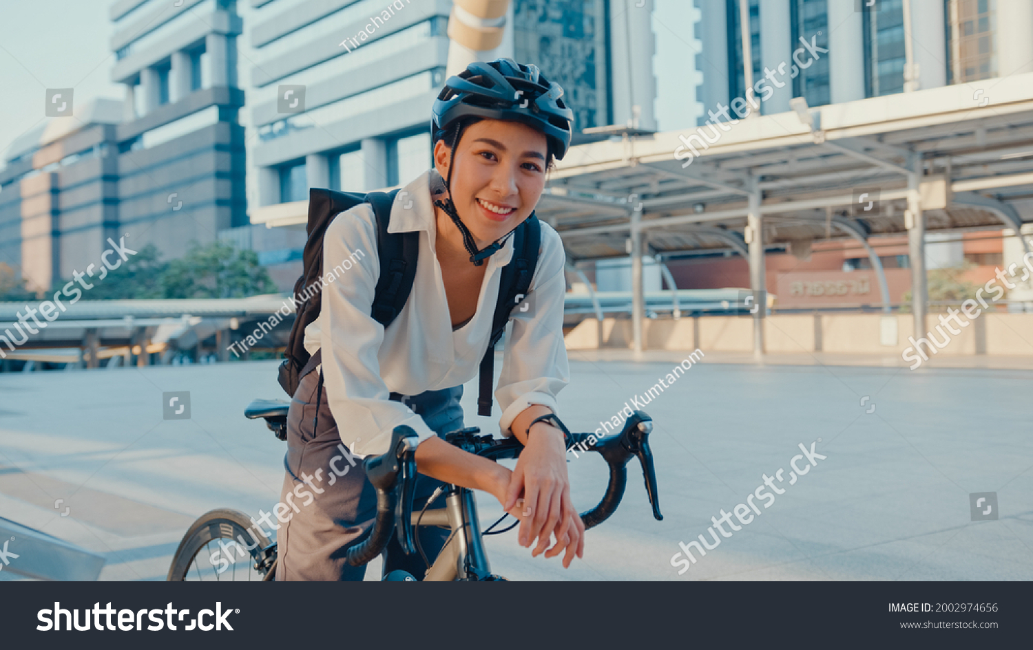 Asian businesswoman go to work at office stand and smiling wear backpack look at camera with bicycle on street around building on a city. Bike commuting, Commute on bike, Business commuter concept. #2002974656