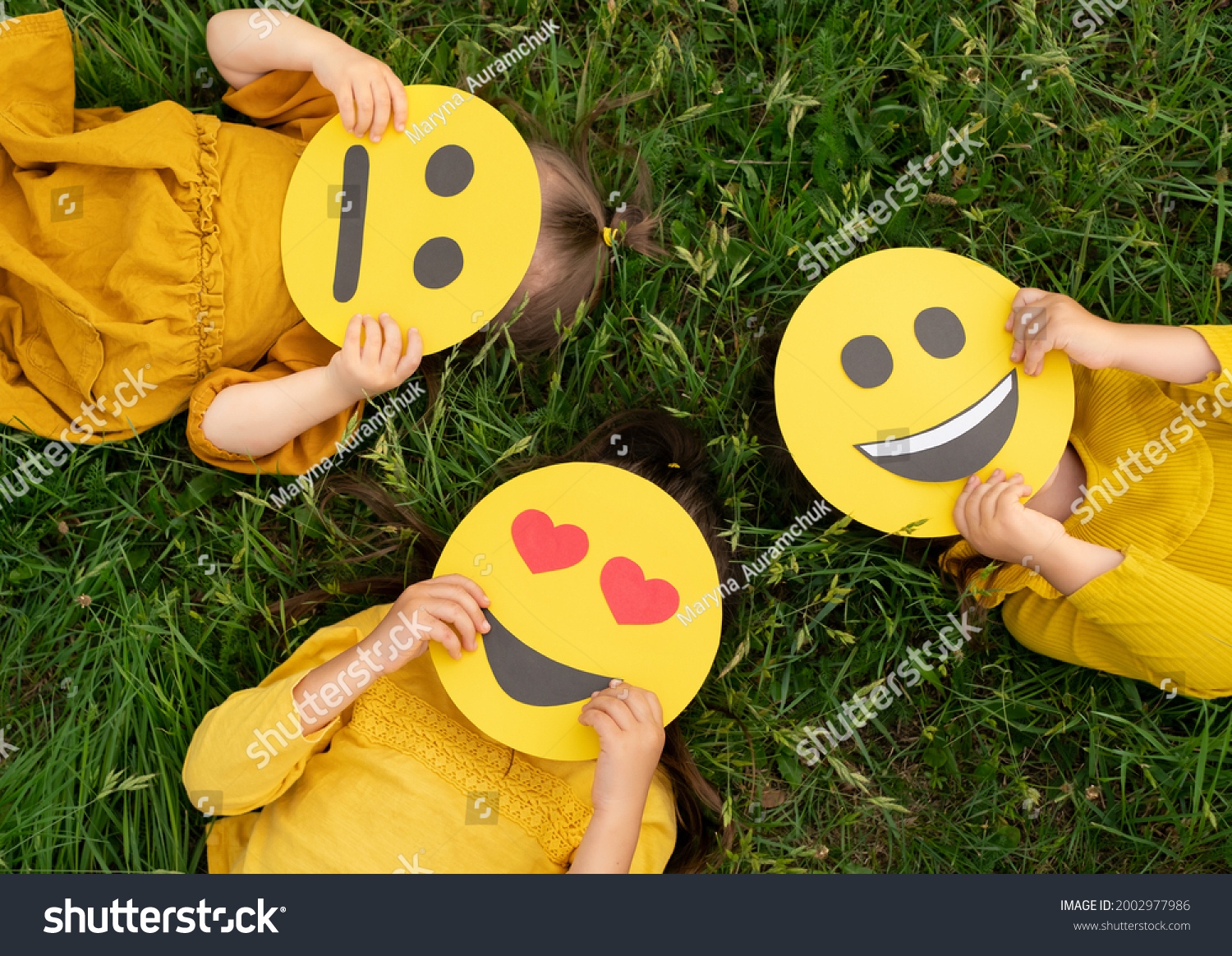 Three children lying on the grass are holding cardboard emoticons with different emotions in their hands: a sad, smiling happy smile, a loving smile with hearts instead of eyes.  World Emoji Day #2002977986