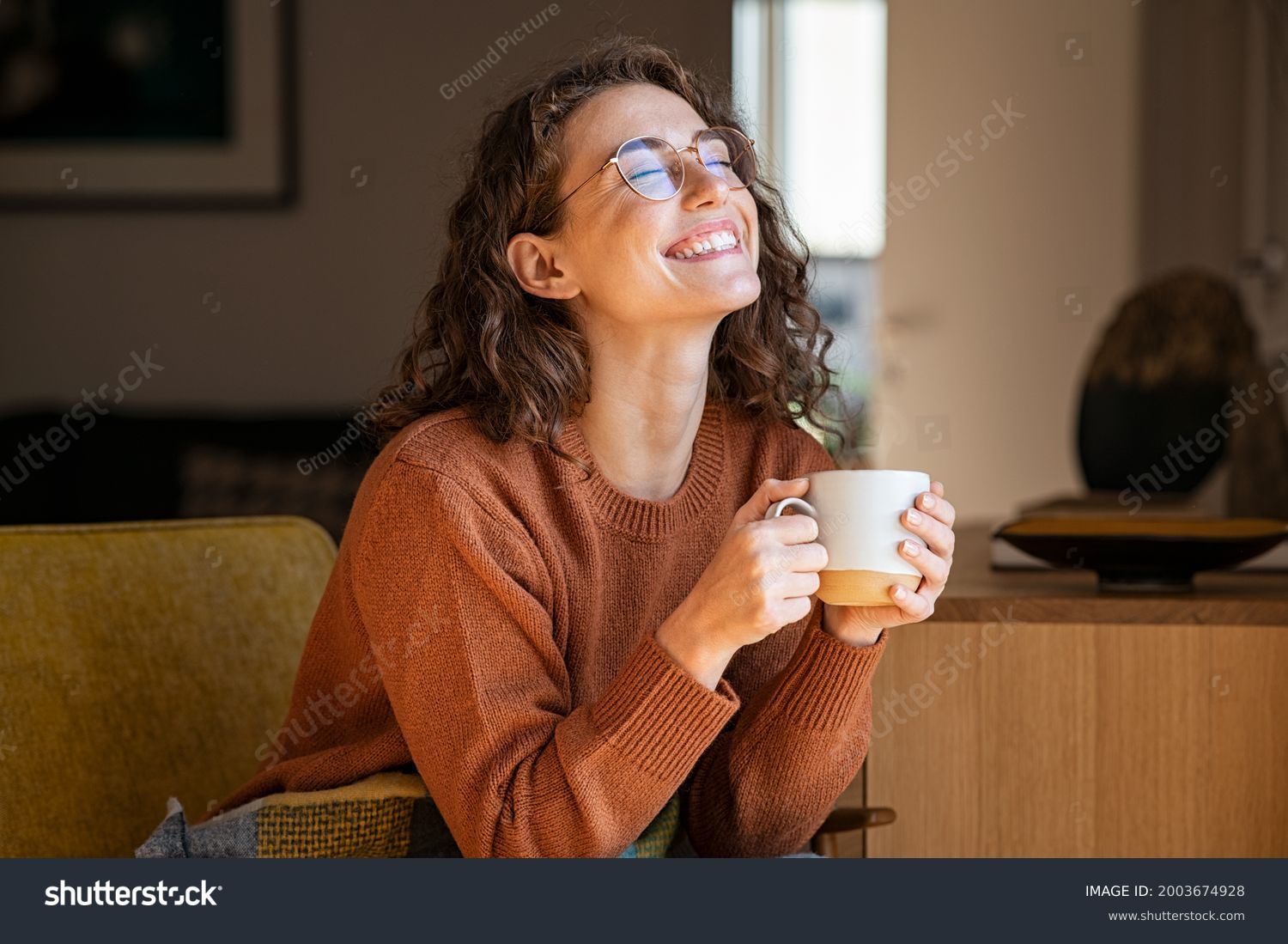 Portrait of joyful young woman enjoying a cup of coffee at home. Smiling pretty girl drinking hot tea in winter. Excited woman wearing spectacles and sweater and laughing in an autumn day. #2003674928