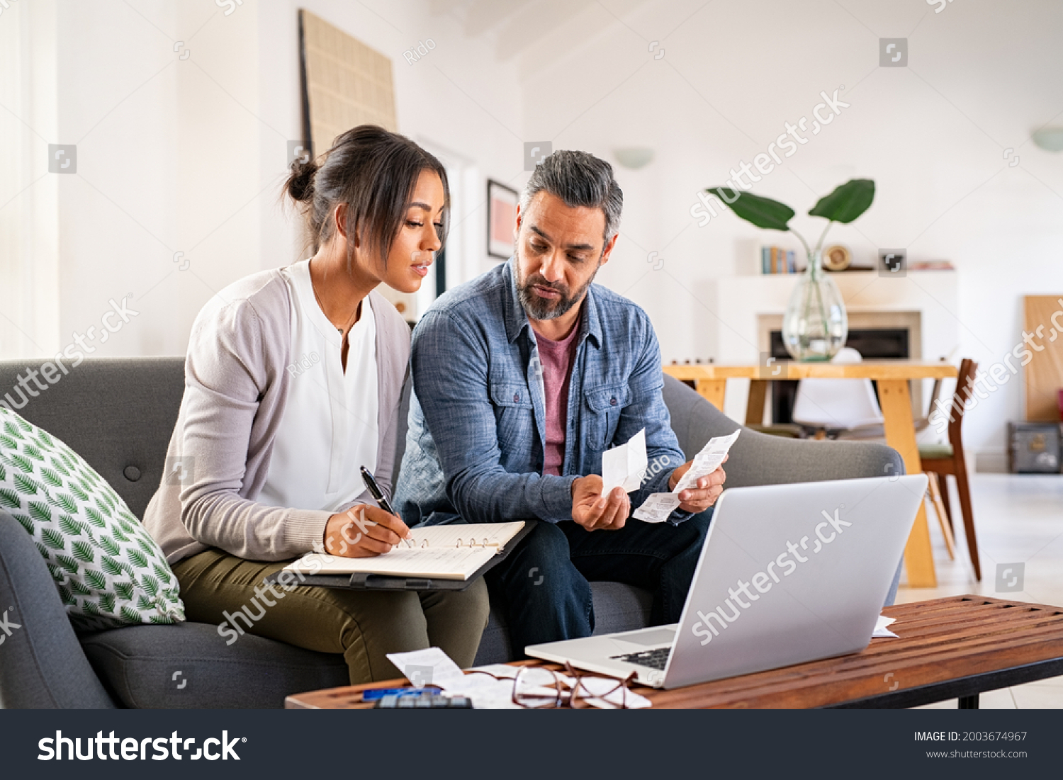Mature couple calculating bills at home using laptop. Multiethnic couple working on computer while calculating finances. Mature indian man with african american woman at home analyzing their finance. #2003674967