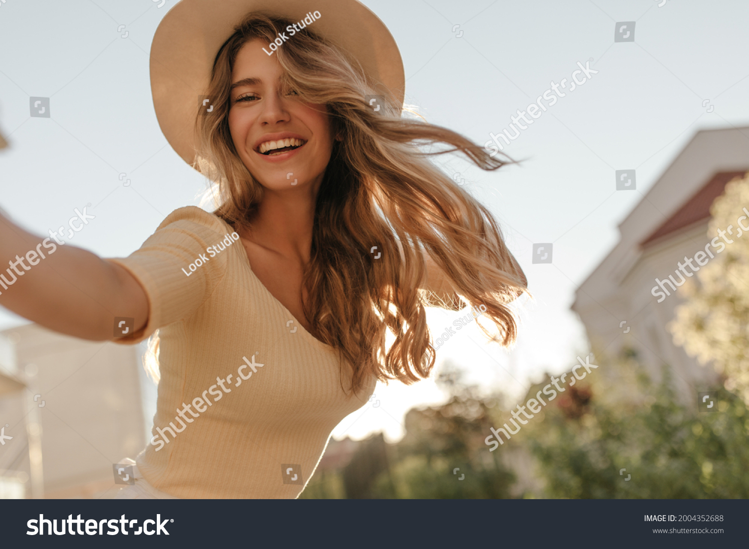 Happy young model makes photo holding camera in hand close-up. Her hair is flying in wind, she is wearing hat. Outdoor photo. #2004352688