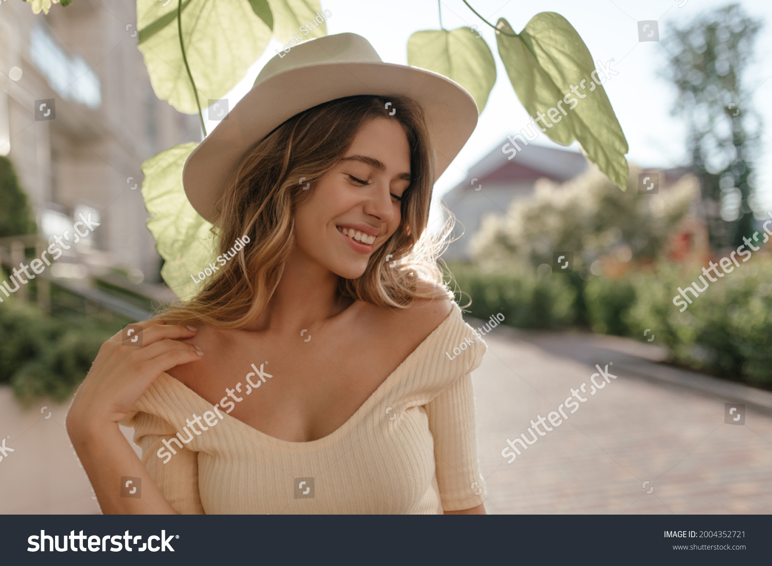 Sunny image of young stylish woman standing on street, in fashionable hat close-up. She has gentle smile and closed eyes. Nice neckline and bare shoulders. #2004352721