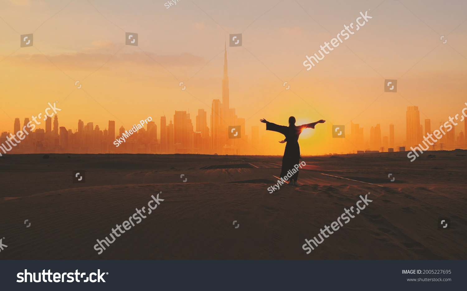 Arabic woman weared in traditional UAE dress - abayain rising her hands on the sunset at a desert with Dubai city silhouette on the background. #2005227695