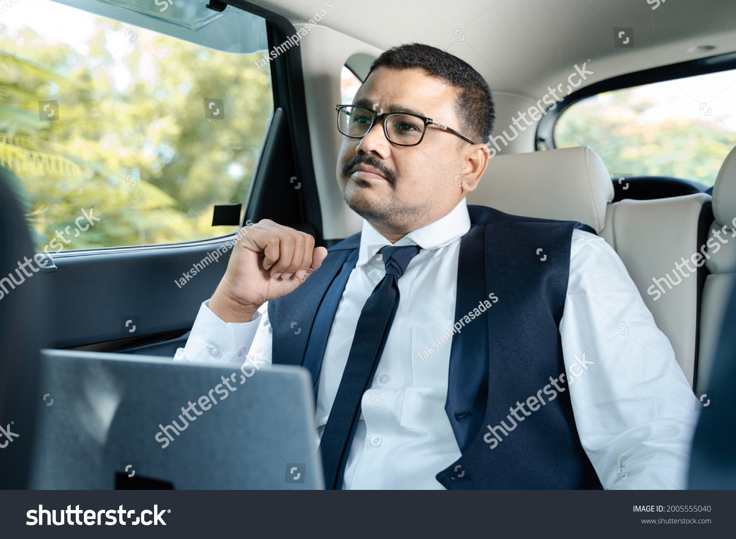 Thinking Serious businessman with laptop travelling in car or cab - Concept of thoughtful and worried about work and family #2005555040
