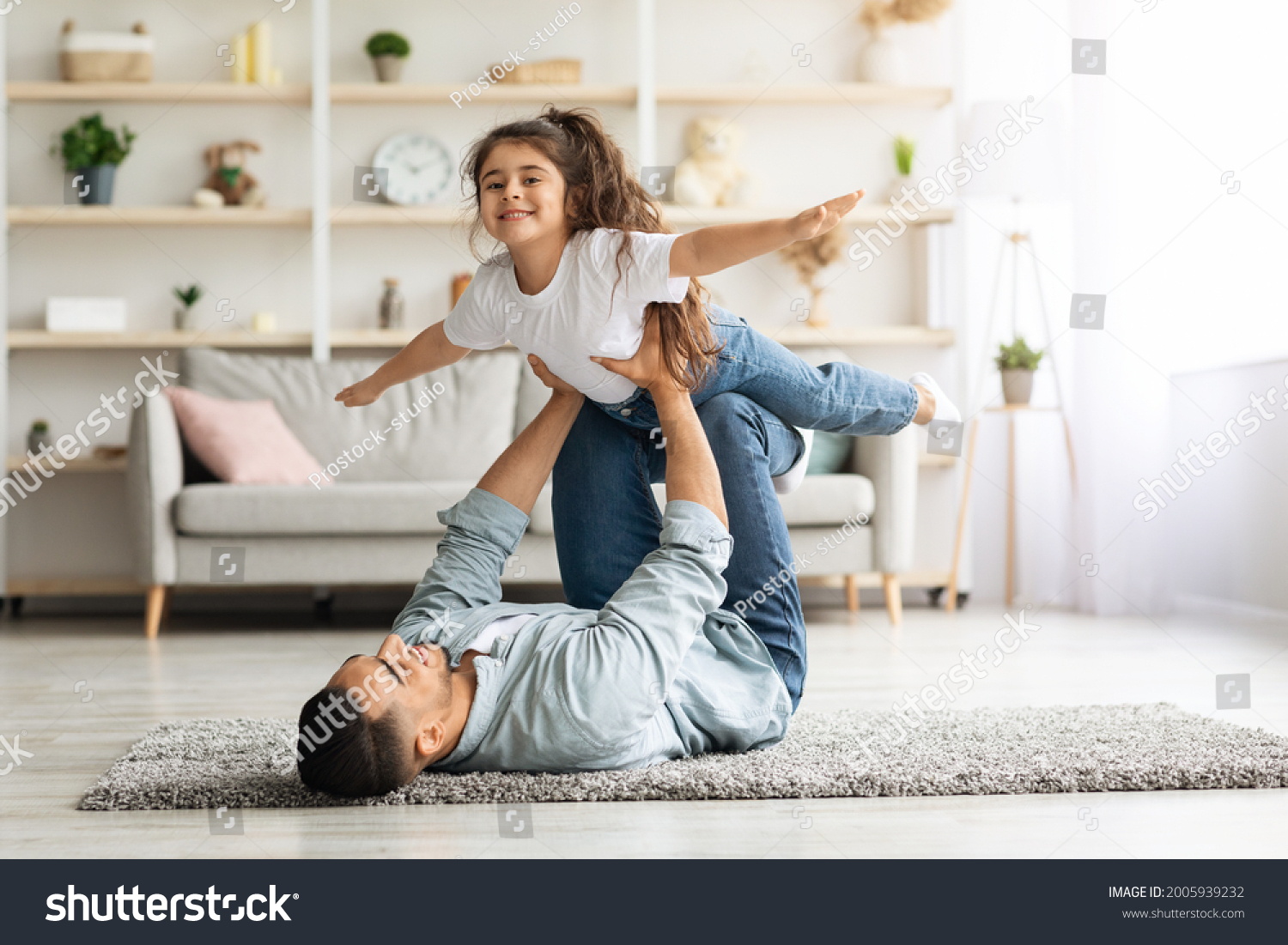 Adorable little curly girl spending time with her daddy at home, middle-eastern young man father playing airplane with his cute daughter, laying on floor in living room, lifting kid up, copy space #2005939232