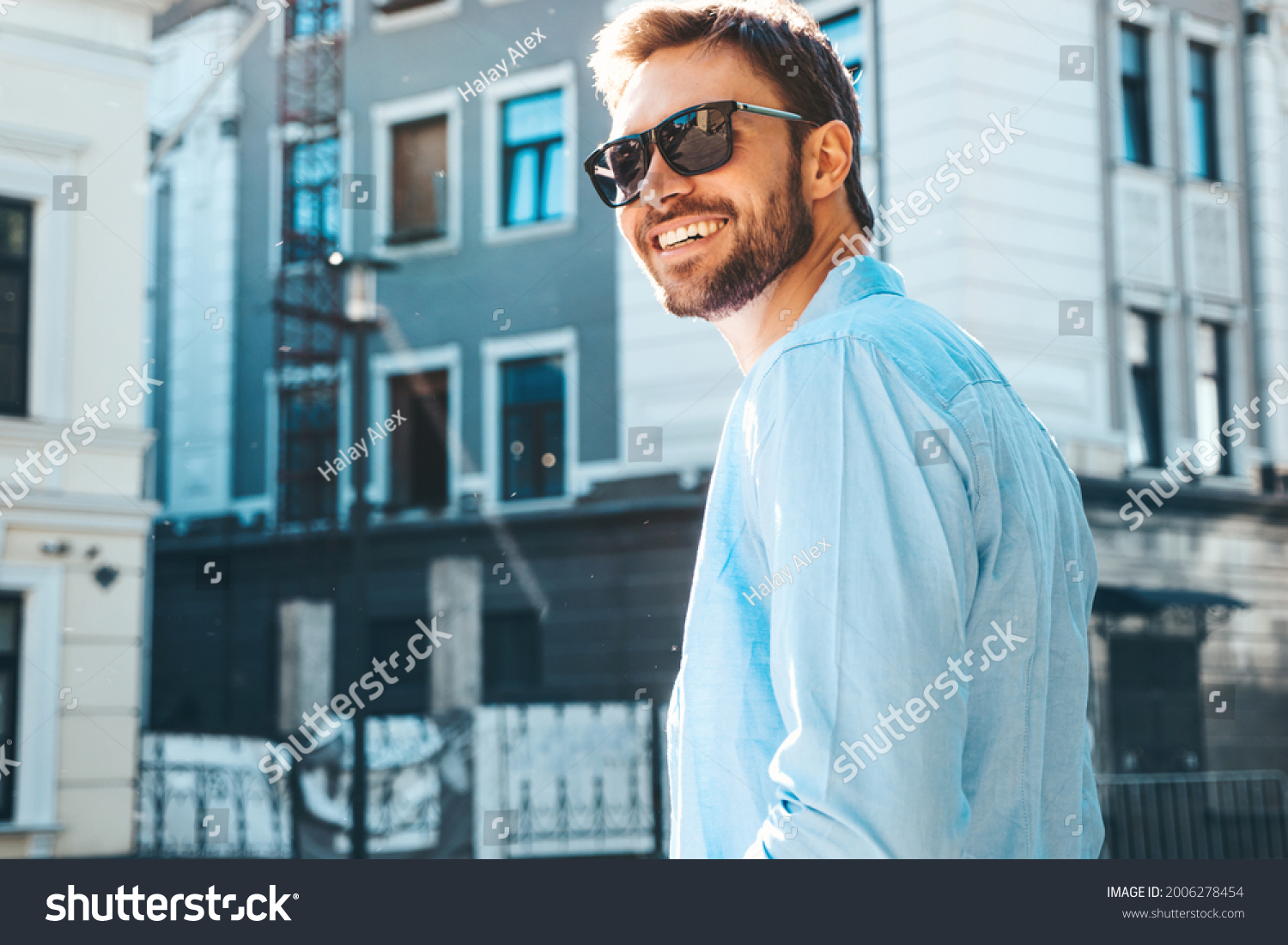 Portrait of handsome smiling stylish hipster lambersexual model.Modern man dressed in blue shirt. Fashion male posing near skyscraper on the street background in sunglasses. Outdoors at sunset  #2006278454