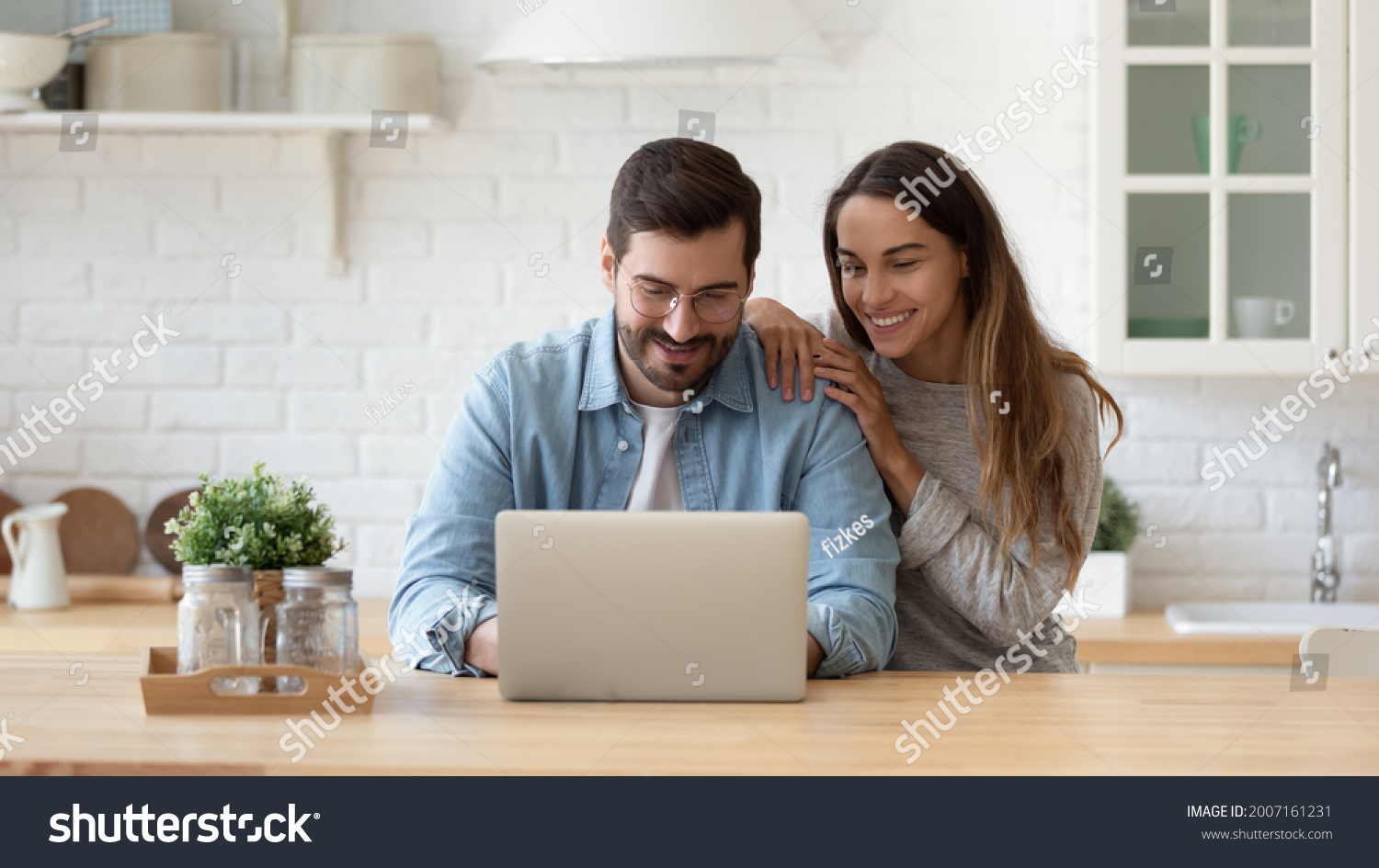 Attractive 35s couple sit at table in cozy kitchen looking at laptop screen, buying goods, tickets online, using e-commerce retail e-services feel satisfied, enjoy comfort usage of modern technology #2007161231