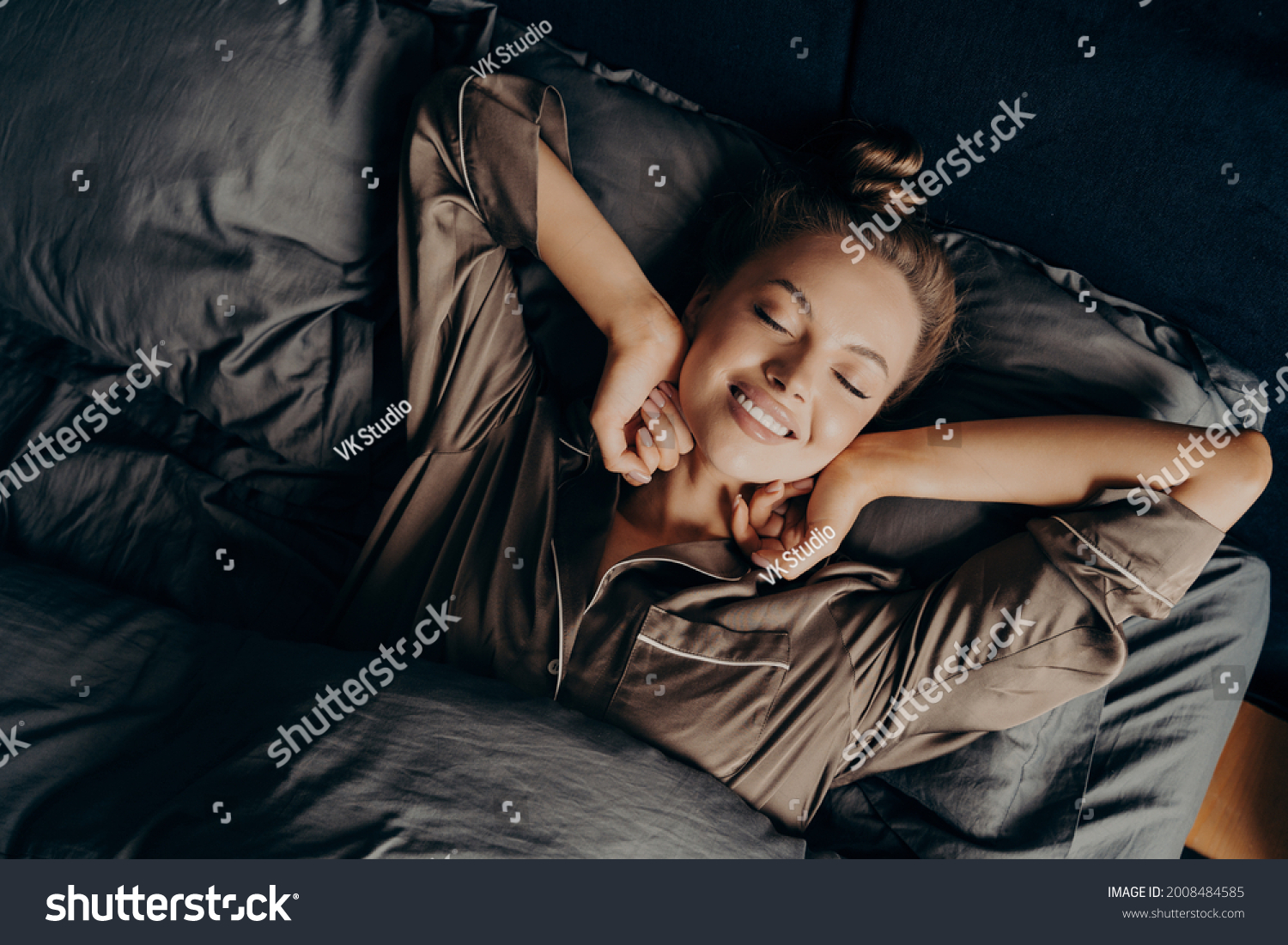 Good morning. View from above of young beautiful relaxed brunette female with closed eyes in satin pajamas waking up in her bed fully rested, happy woman stretching after night sleep #2008484585
