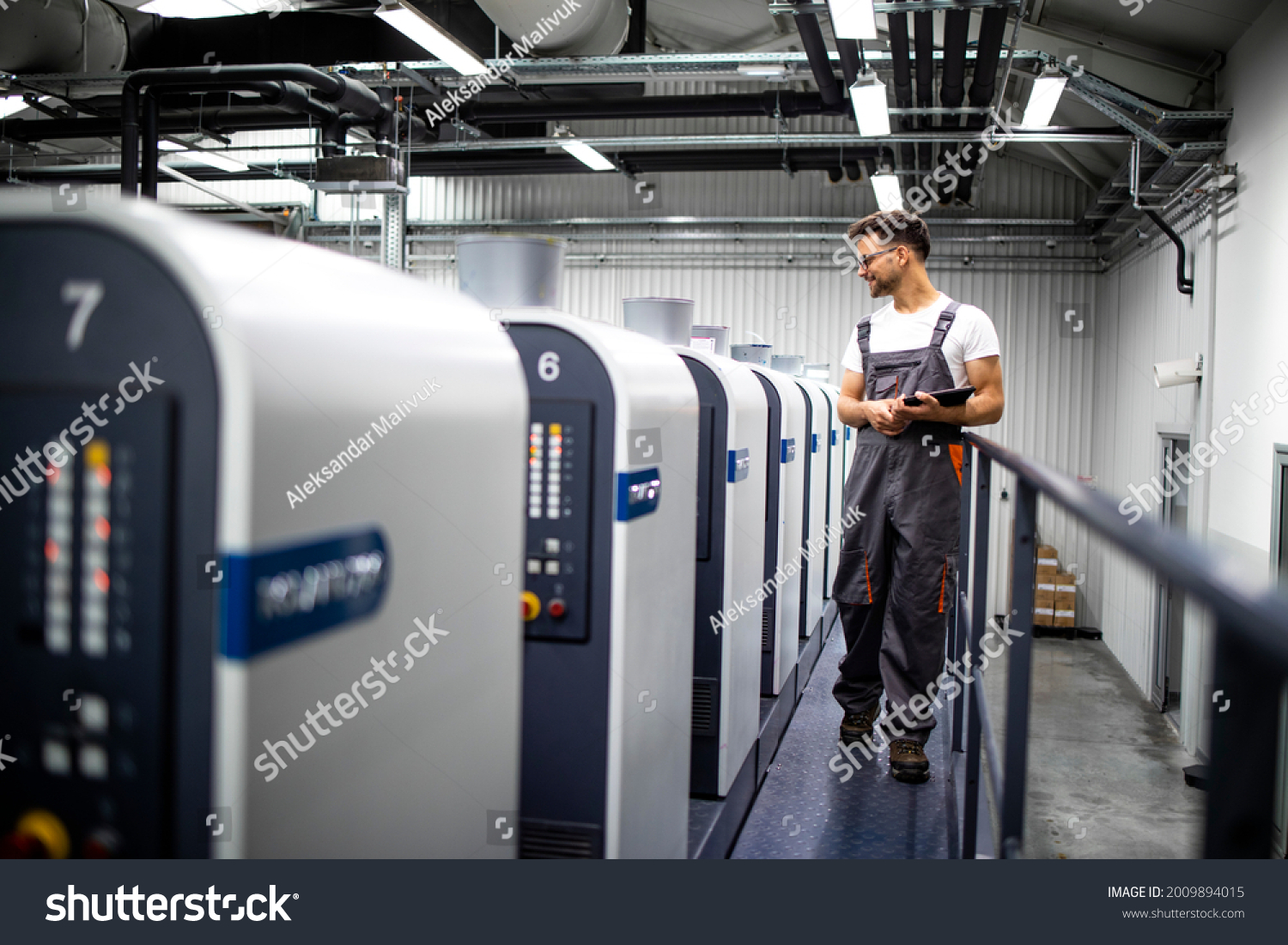 Printing house interior with modern offset print machine and operator in working uniform checking quality and controlling process of print. #2009894015