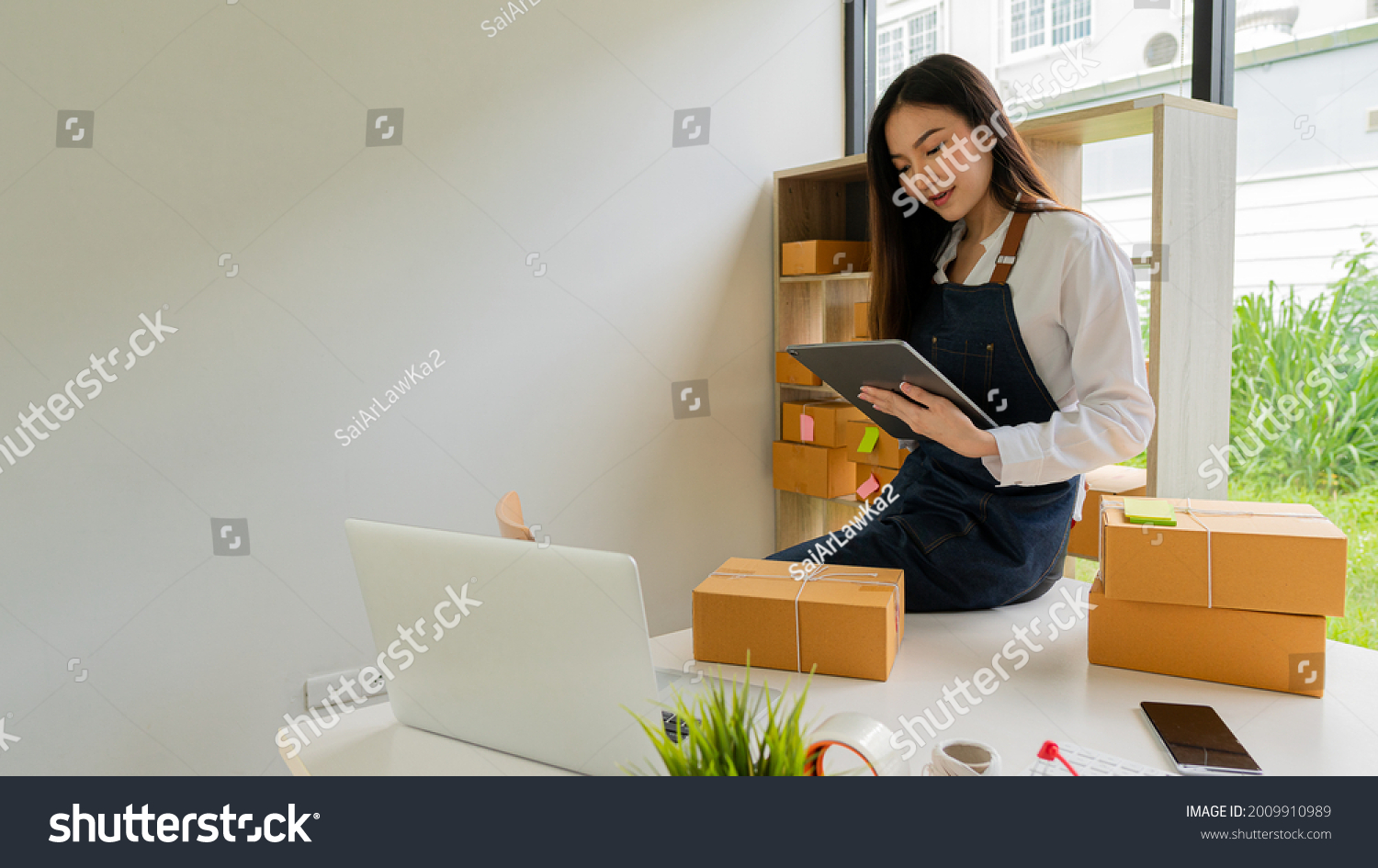 Independent Asian woman SME business owner online shopping working on laptop computer with parcel box on table at home - online SME business and online shipping and shopping concept. #2009910989