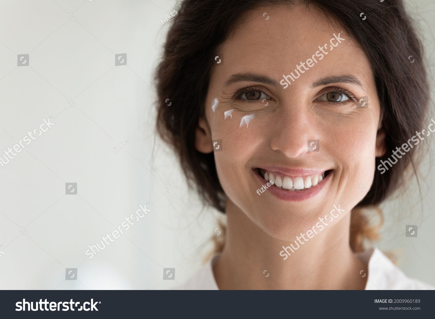 Close up portrait of smiling young latin female face with cosmetic cream moisturizer drops on skin under eyes. Happy millennial lady look at camera introduce new antiage creme serum lotion. Copy space #2009960189