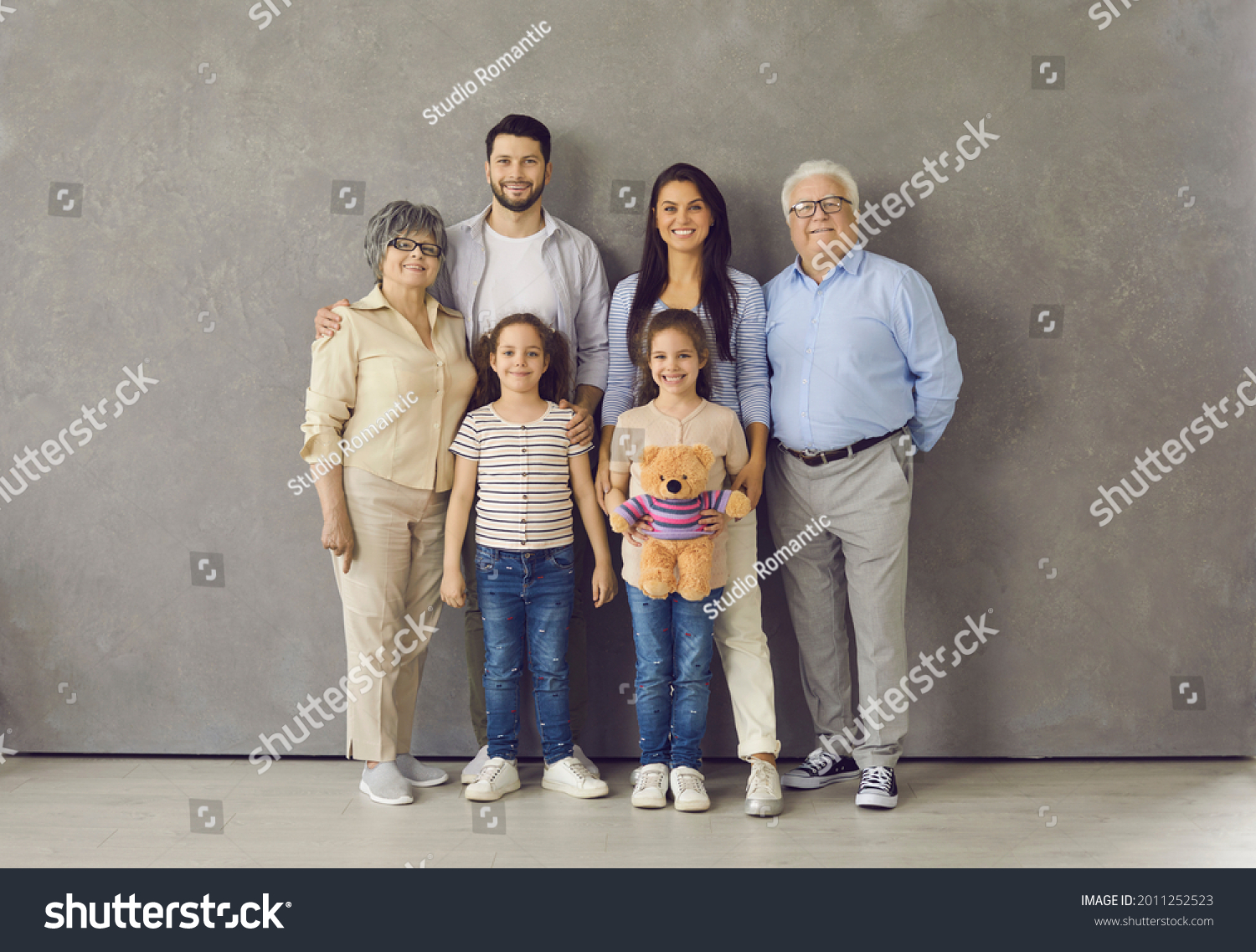 Studio photoshoot group portrait of happy big extended multi generational family. Cheerful mom, dad, grandma, grandpa and two little daughters with toy standing together, looking at camera and smiling #2011252523