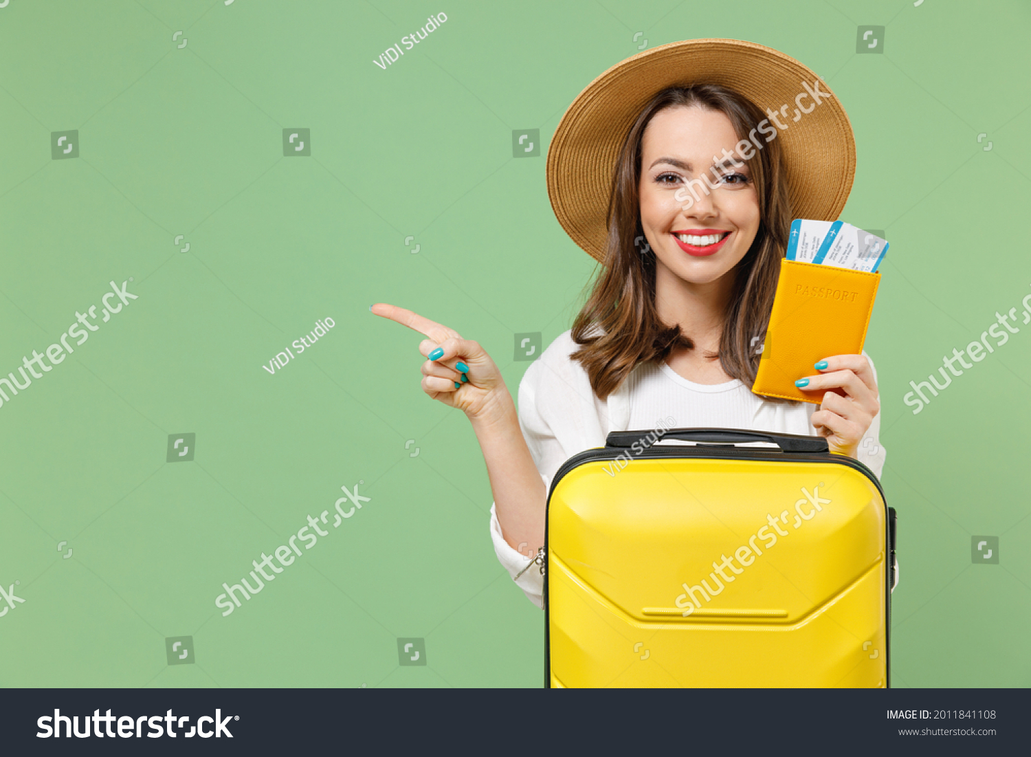 Close up tourist woman in casual clothes hat hold passport tickets yellow suitcase point finger aside workspace isolated on green background Passenger travel abroad weekend getaway Air flight concept. #2011841108