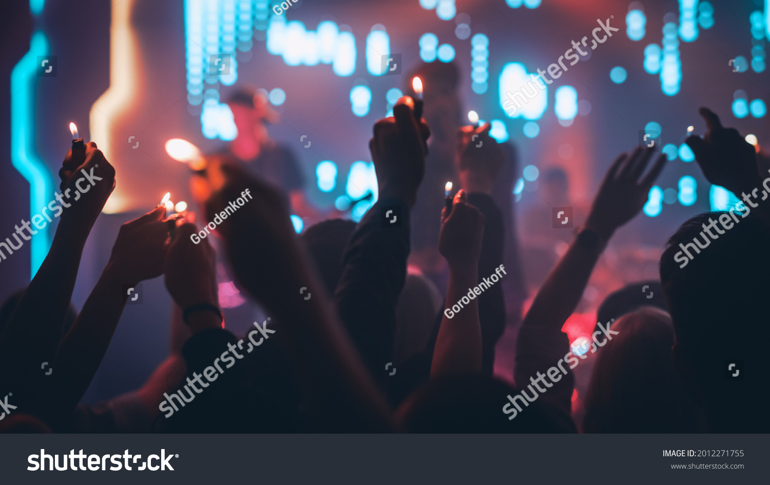 Rock Band Performing a Slow Song at a Concert in a Night Club. Front Row Crowd is Holding Lighters. Silhouettes of Fans Raise Hands in Front of Bright Colorful Strobing Lights on Stage. #2012271755
