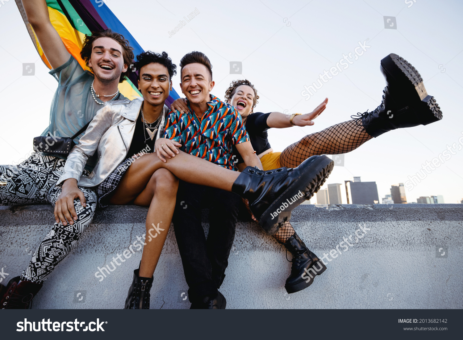 Four LGBTQ people celebrating pride while sitting together. Four friends smiling cheerfully while raising the rainbow pride flag. Group of young queer individuals celebrating together outdoors. #2013682142