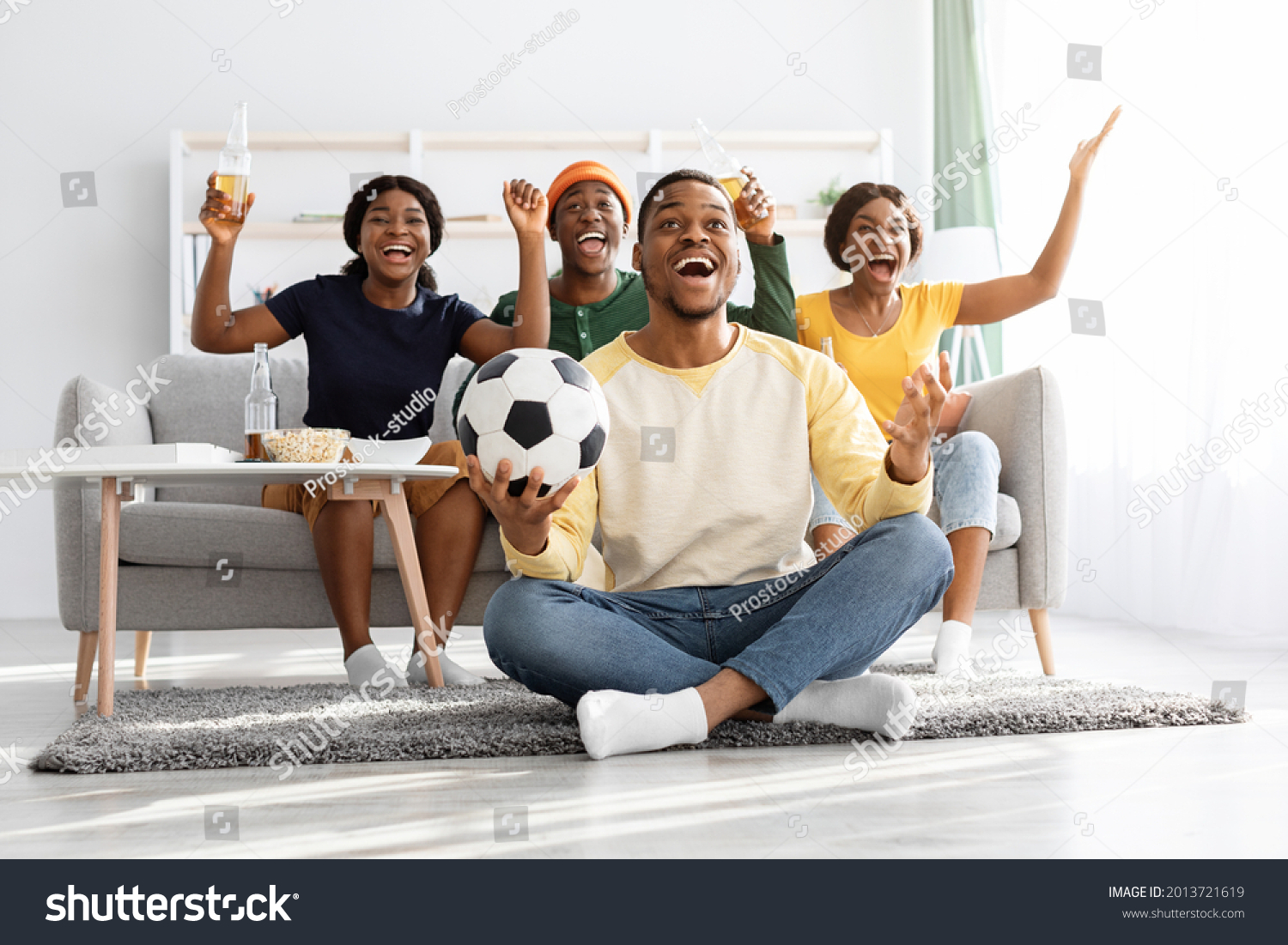 Emotional football fans young african american men and women watching game together at home, celebrating win of their team, raising hands up and screaming, excited black guy with soccer ball #2013721619